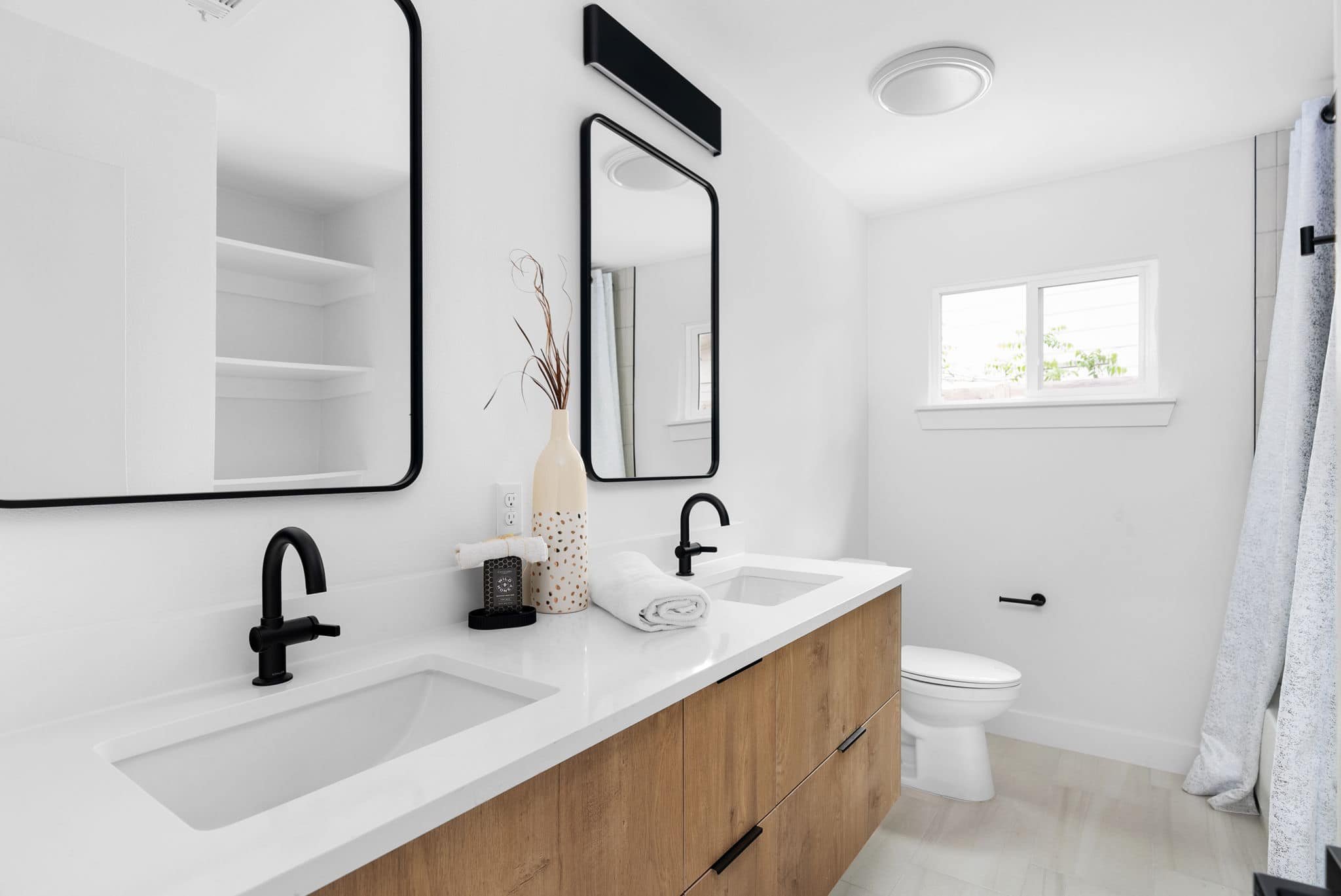 Modern bathroom with double white vanity, black faucets, large mirrors, a small window, a toilet, and a beige wooden cabinet, decorated with a tall vase with dried plants.