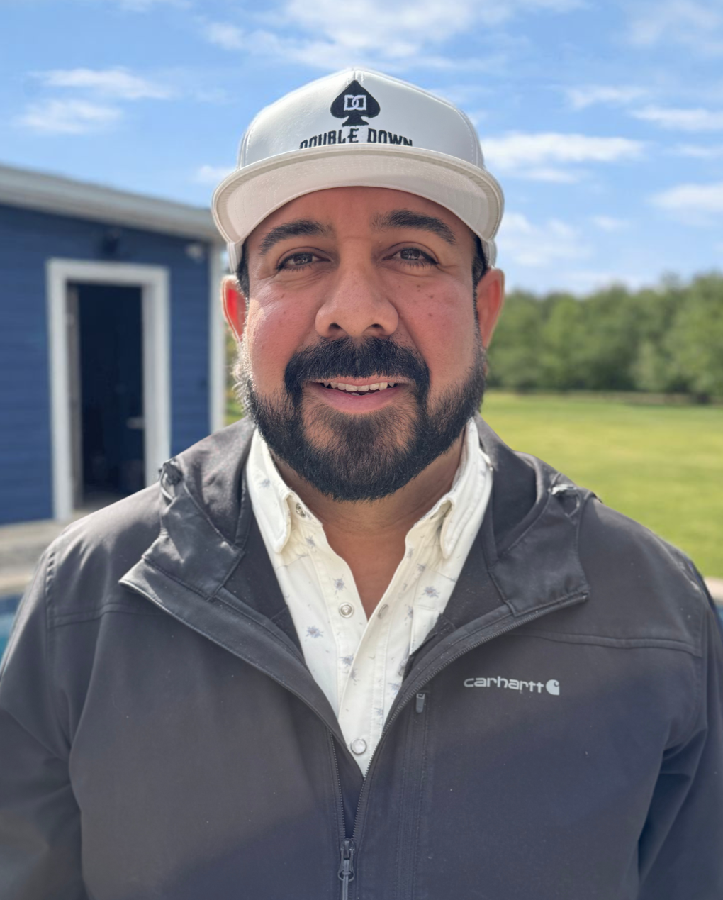 A man with a beard and mustache wearing a white cap with black logo and a black Carhartt jacket, smiling outdoors with a blue building and grassy field in the background.