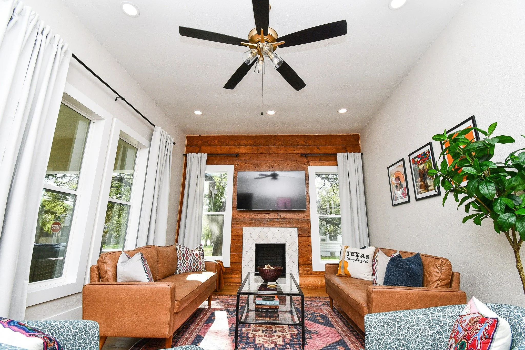 Living room with two brown leather sofas, decorative pillows, a glass coffee table, large windows with white curtains, a flat-screen TV above a fireplace, wood-paneled wall, framed pictures on the white wall, a ceiling fan, and a potted plant.