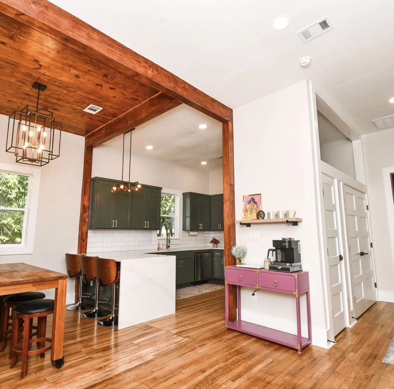 Open-concept kitchen and dining area with wooden floors, dark green cabinets, and a pink side table with a coffee machine on top.