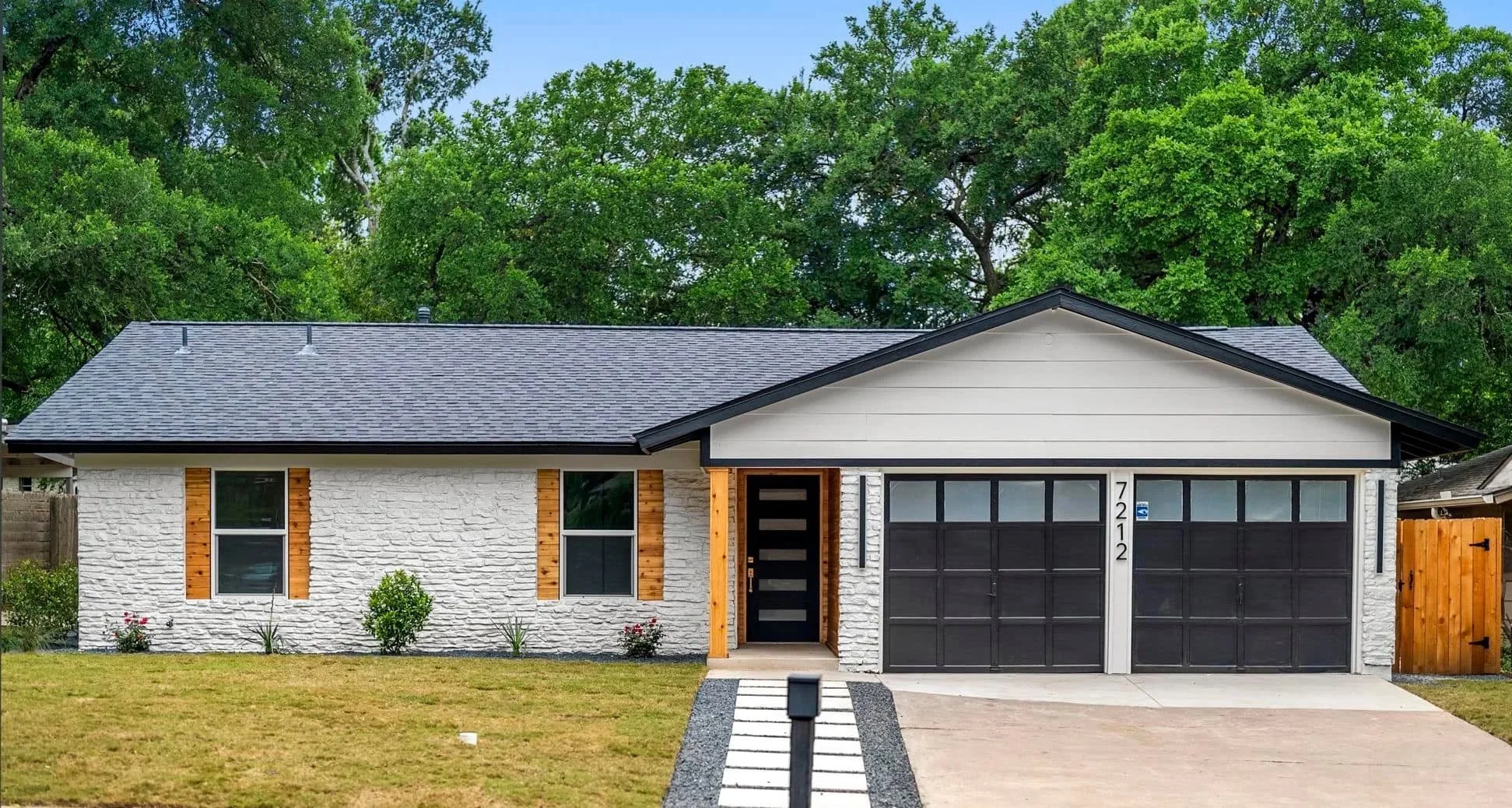 Modern single-story house with white brick and wooden accents, black garage doors, and a well-maintained front yard with a sidewalk leading to the front door, set against a backdrop of lush green trees.
