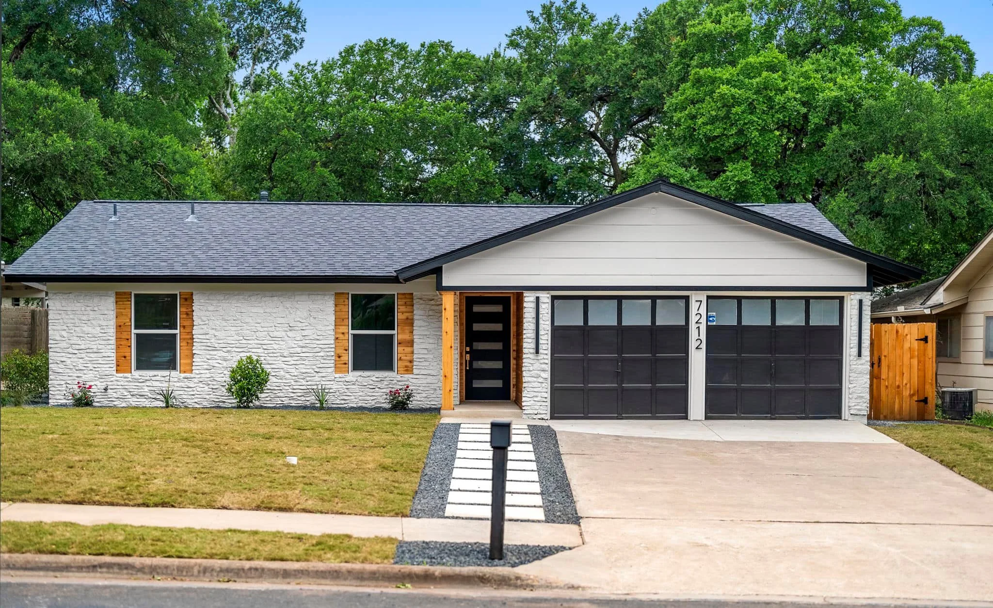 A modern single-story house with a white brick exterior, black garage doors, and a black front door with horizontal glass panels. The house features wooden accents around the windows and a manicured lawn in front. Tall trees are visible in the backgr
