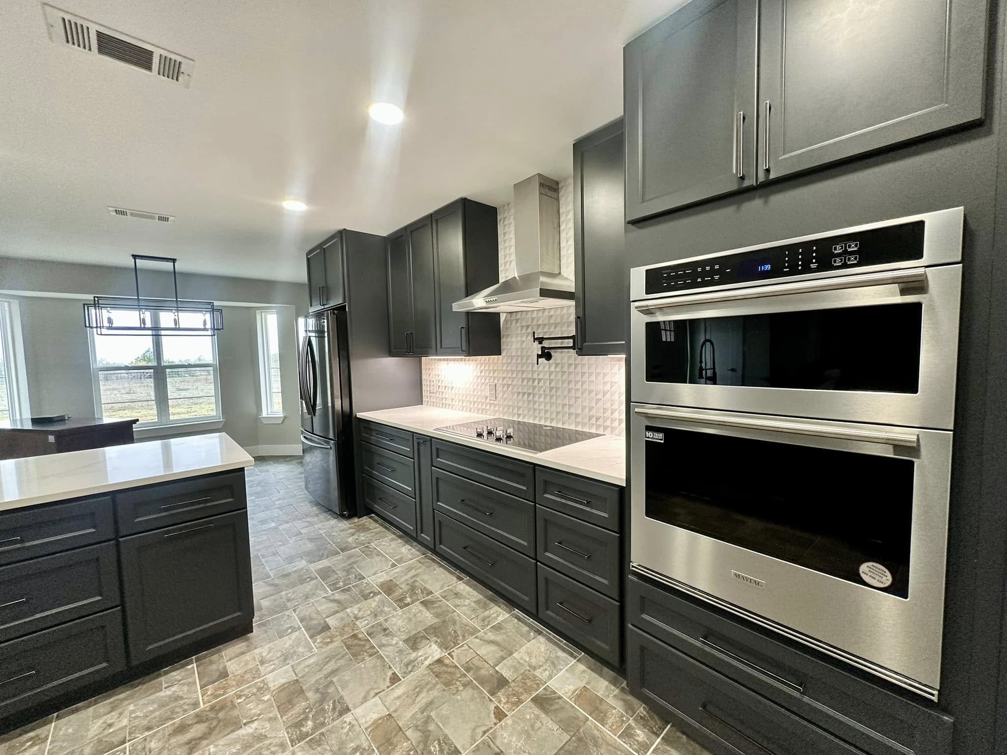 Modern kitchen with dark gray cabinets, stainless steel double oven, fridge, white countertops, textured white backsplash, and tiled floor.