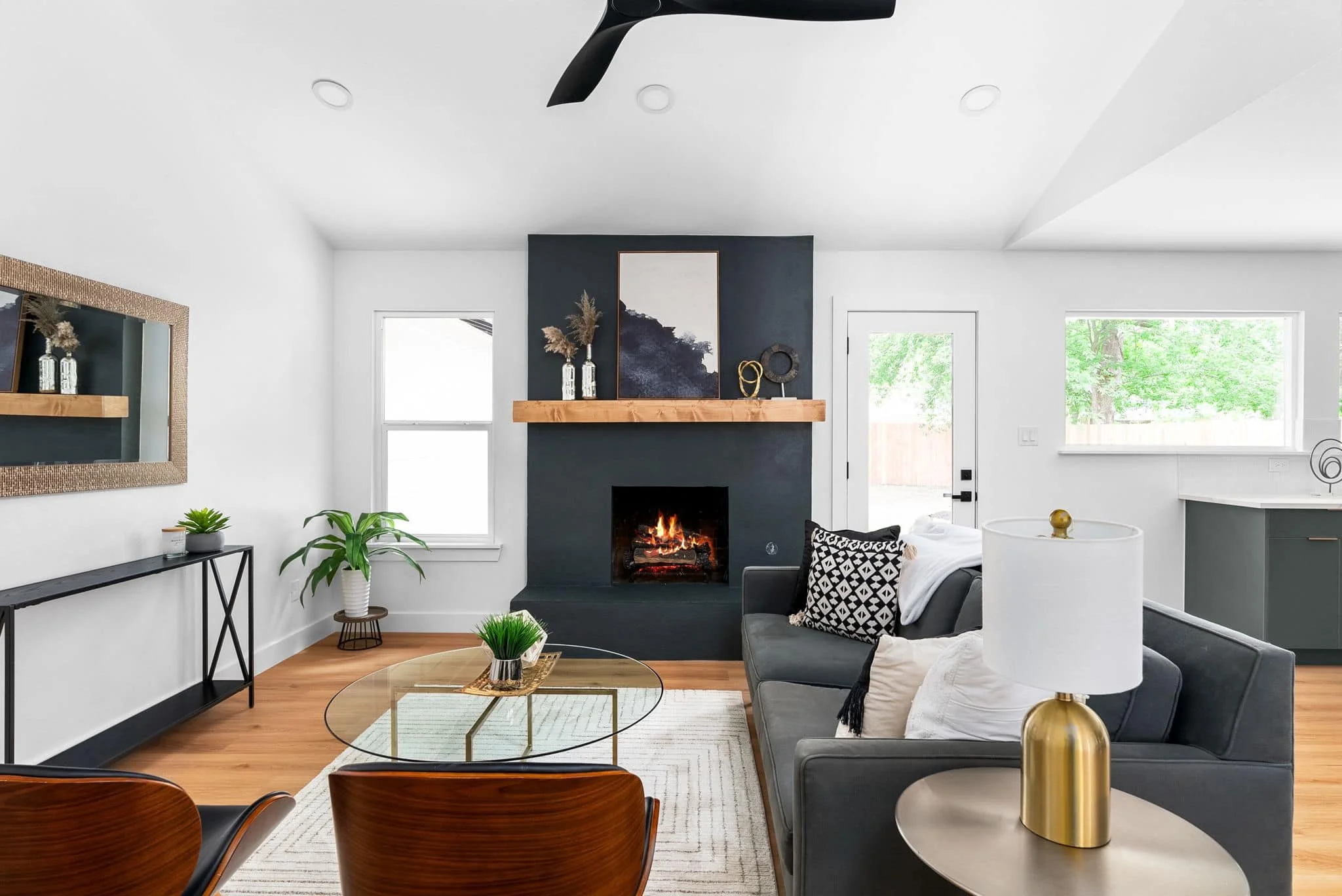 Living room with a white wall, black accent wall with a fireplace, wooden mantel, and decorated with art and sculptures. There are large windows, a grey sofa with black and white cushions, a glass top coffee table with a plant, a black console table 