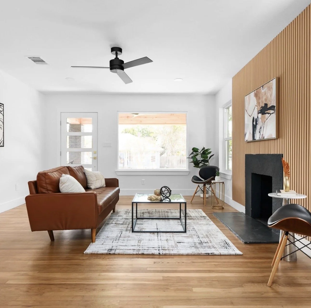 Living room with a brown leather sofa, a black and wood chair, a glass coffee table, a fireplace with a black surround, a large window, and a wooden accent wall with artwork. Light wood flooring, white walls, and a ceiling fan.