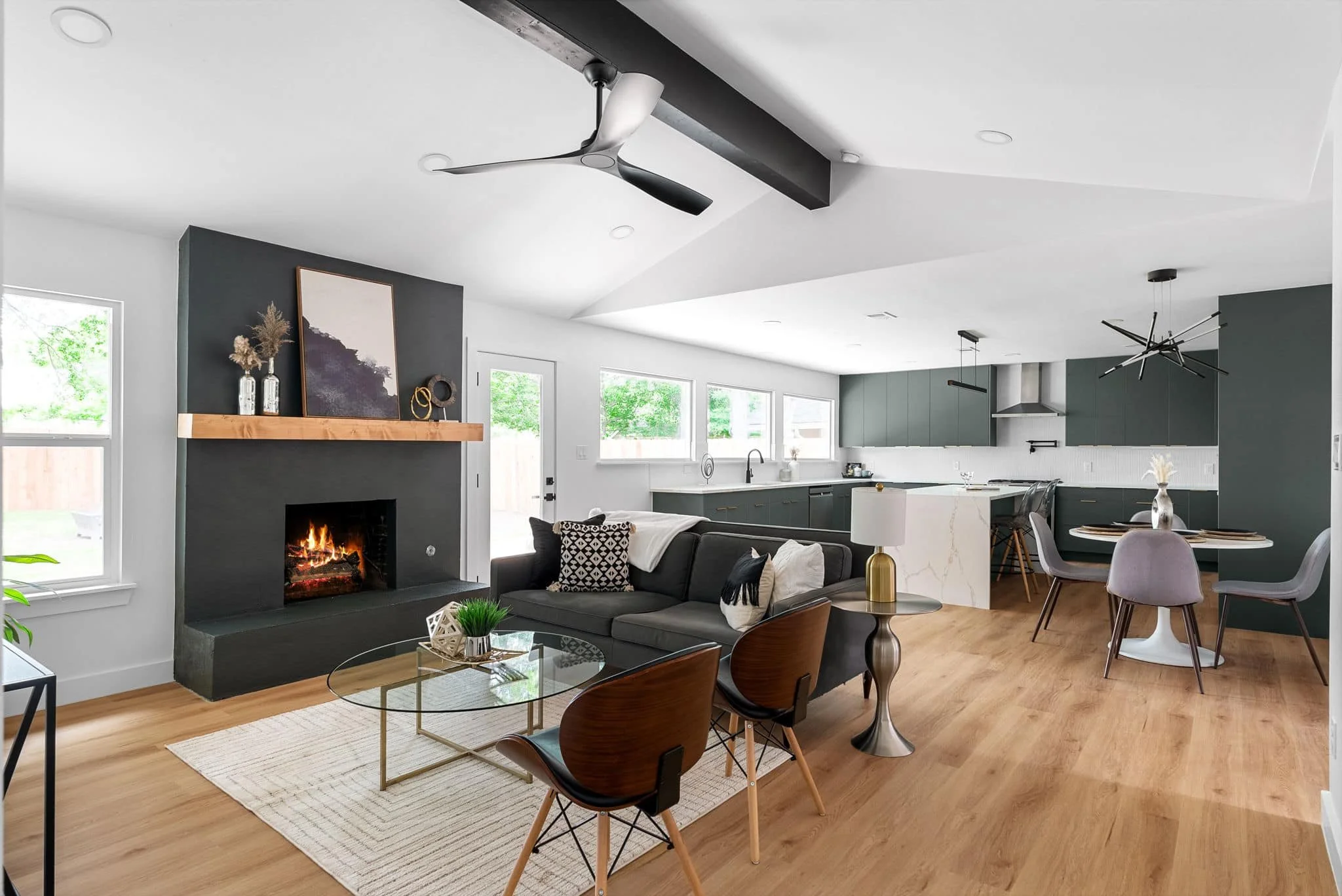 Open-concept living room and kitchen with modern decor, wood flooring, gray and white walls, a black fireplace with fire, a ceiling fan, and contemporary furniture.