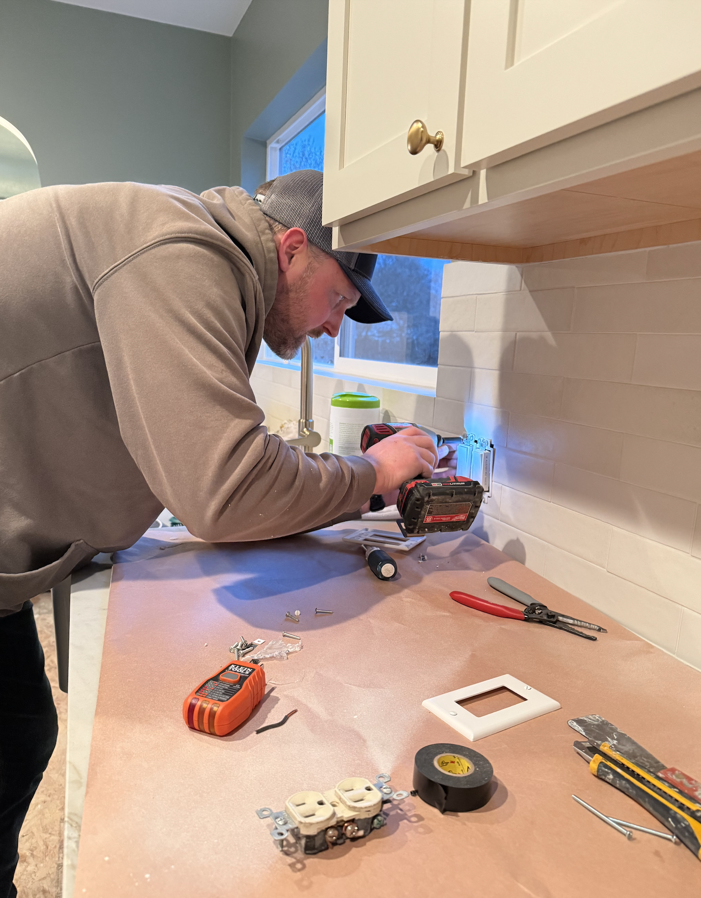 Man installing or repairing electrical outlets in a kitchen, with tools and electrical components on the countertop.