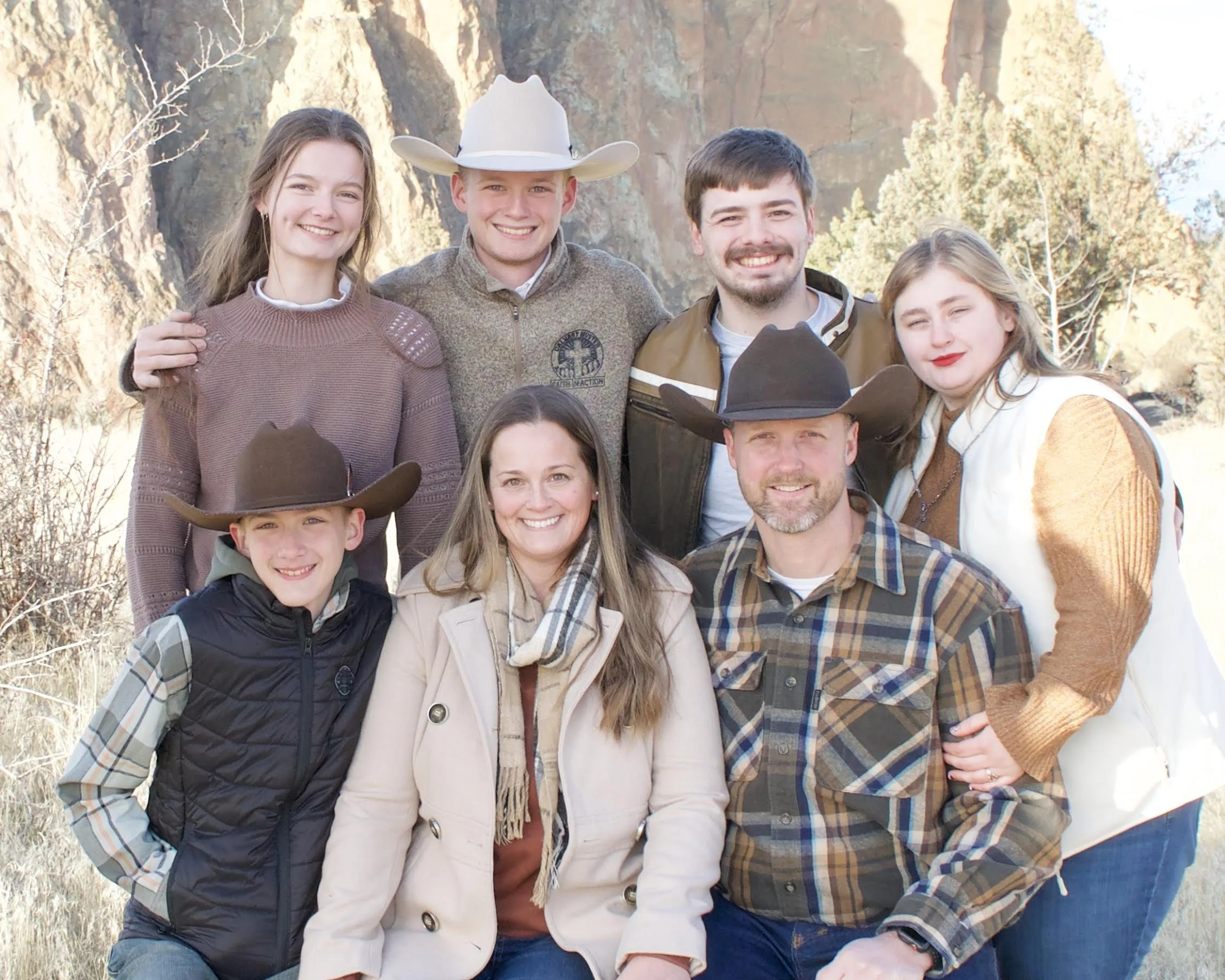 A family of nine in outdoor setting with rocky hills and trees, dressed in casual and cowboy attire, smiling for a group photo.