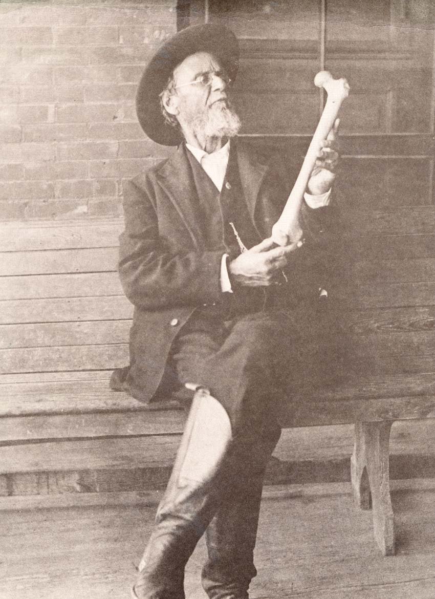 A. T. Still, wearing glasses, a dark hat, and a suit, sits on a wooden bench holding a large bone. Based on an article about osteopathy.