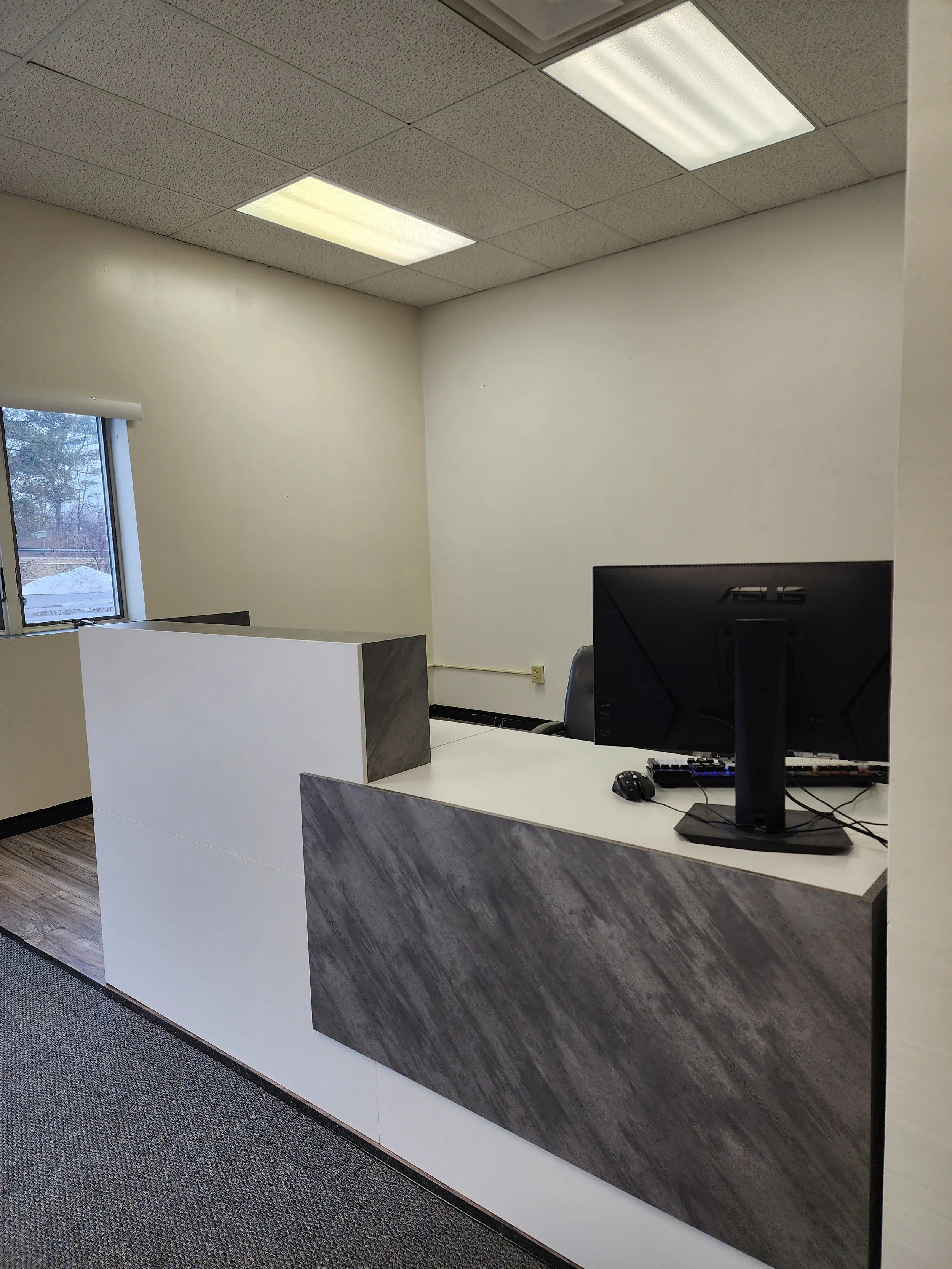An office reception area with a modern white and gray desk and a computer monitor. There is a window on the left side showing an outside scene. The ceiling has white acoustic tiles with two fluorescent light panels. The floor is covered with a patterned carpet.