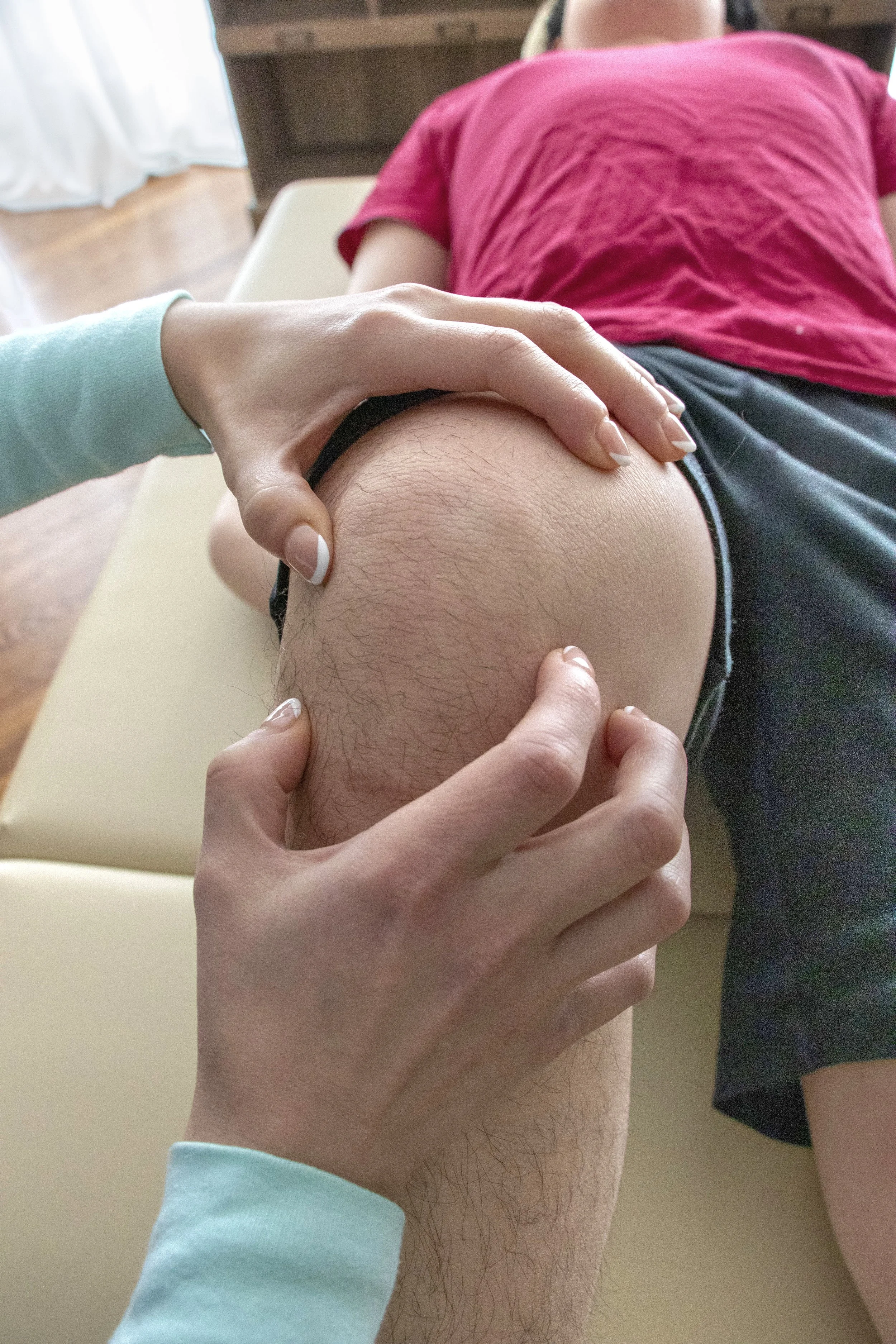 Person lying face down on a padded surface receiving physical therapy or massage on their knee, with a doctor's hands holding and applying pressure to the knee.
