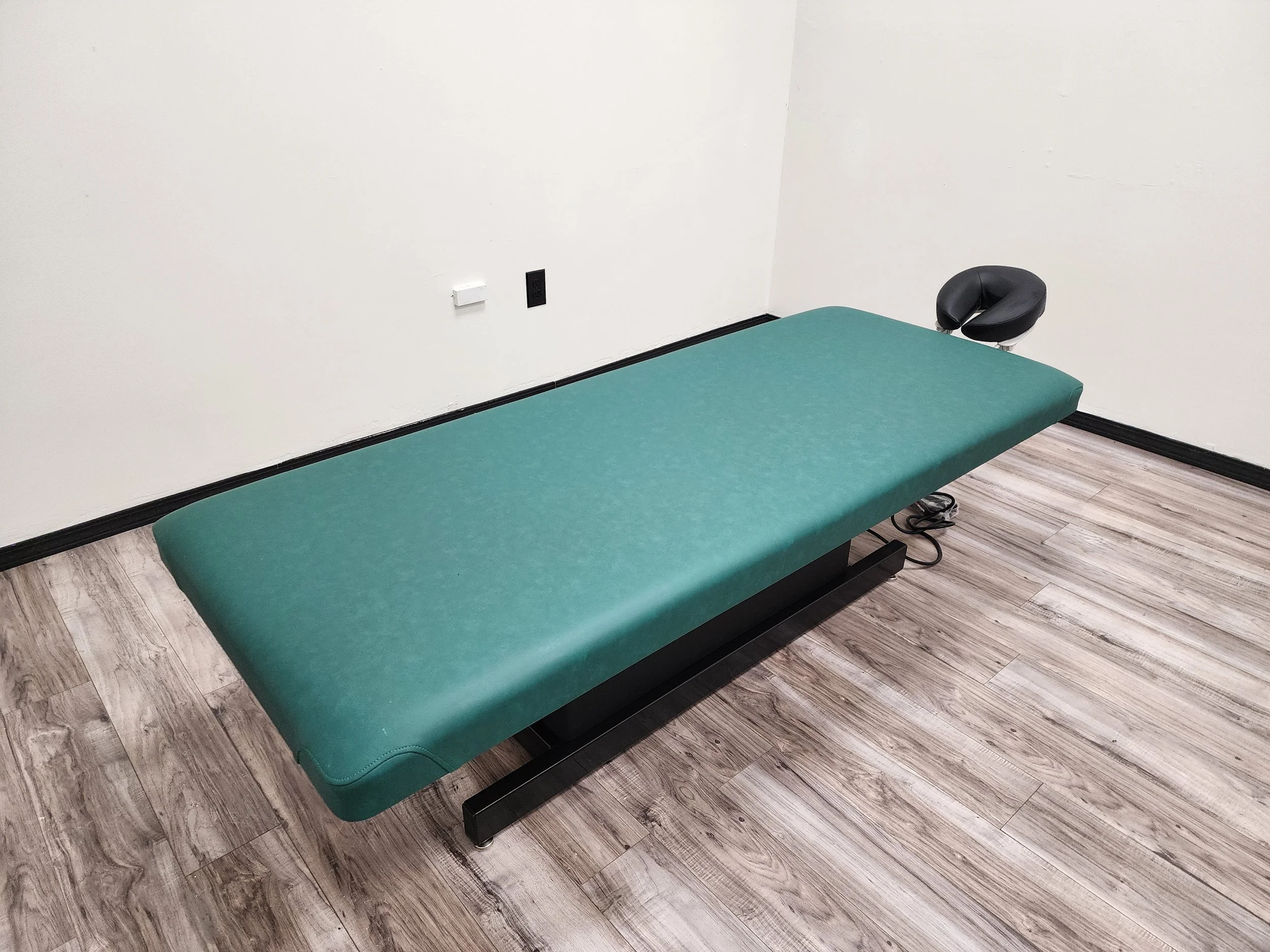 Exam or treatment table with a green cushion in a room with wood-patterned flooring and a plain white wall.