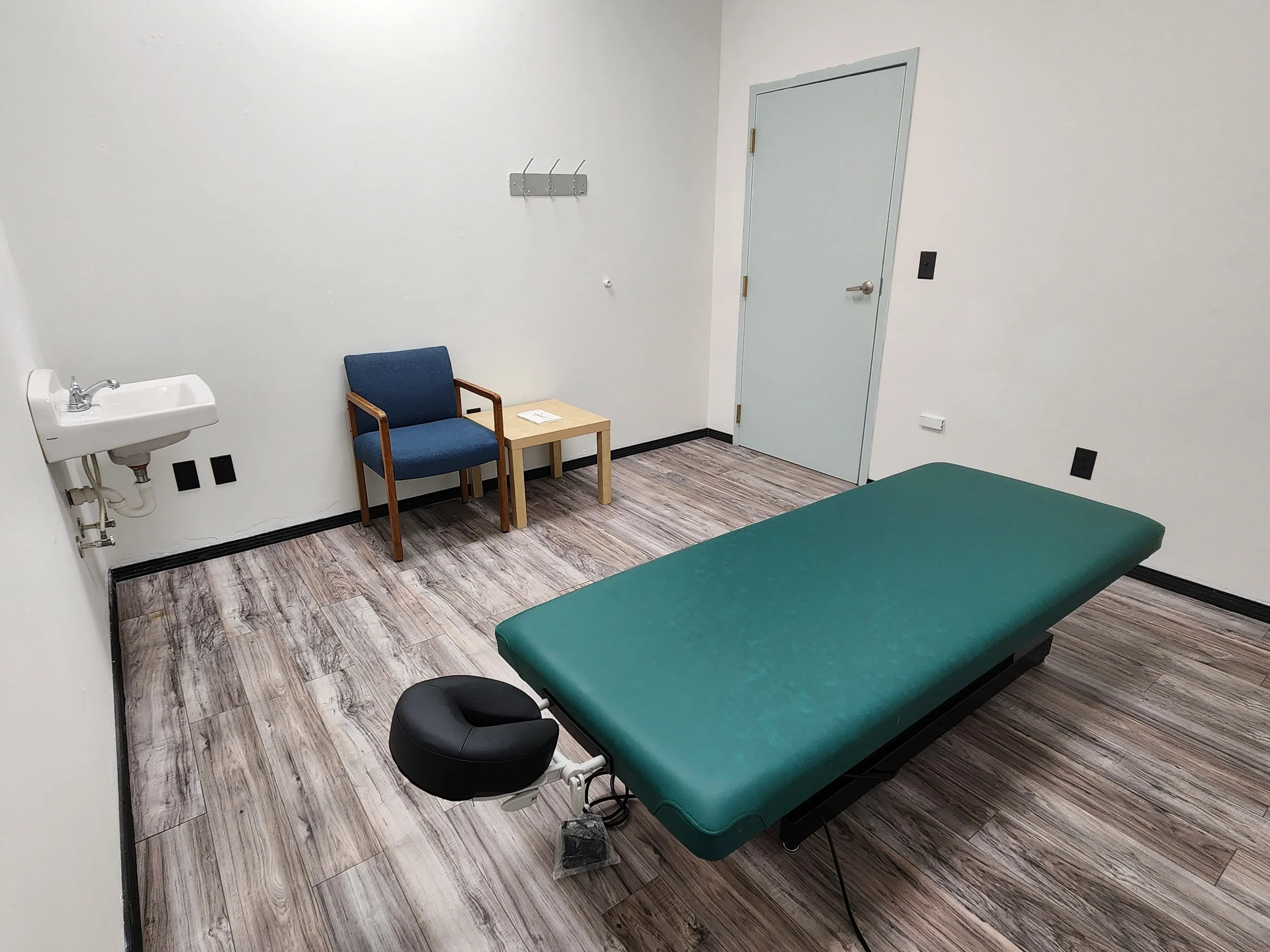 A medical examination room with a green examination table, a black stool, a wall-mounted white sink, a blue chair, a small wooden table, coat hooks on the wall, a closed door, and wood-patterned flooring.