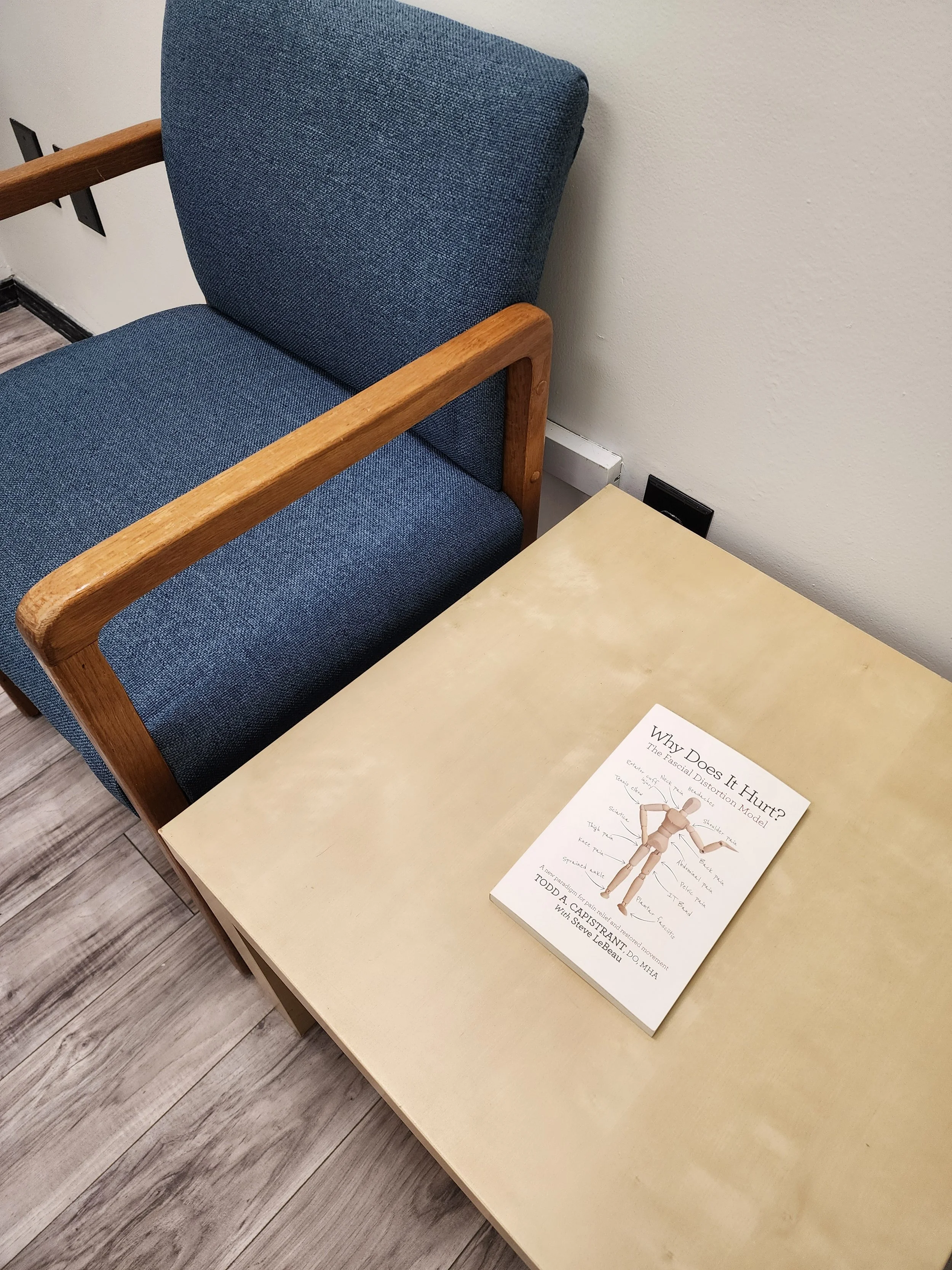A beige table with a book titled 'Why Does It Hurt? The Fascial Distortion Model' placed on it, next to a blue upholstered chair with wooden armrests, in a room with light-colored walls and wood-patterned flooring.