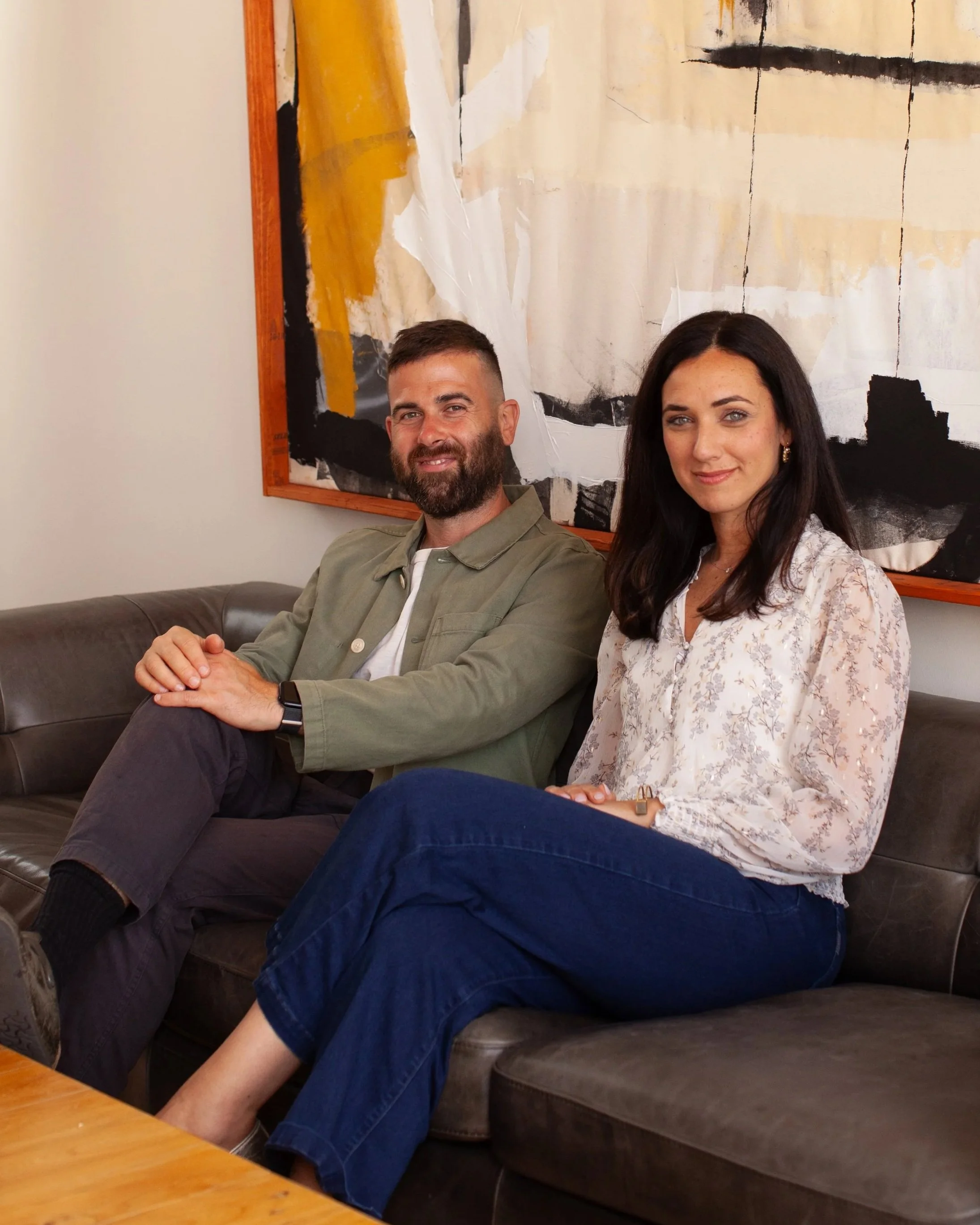 A man and woman sitting on a brown leather couch in front of a modern abstract painting with yellow and black colors, in a room with light-colored walls.