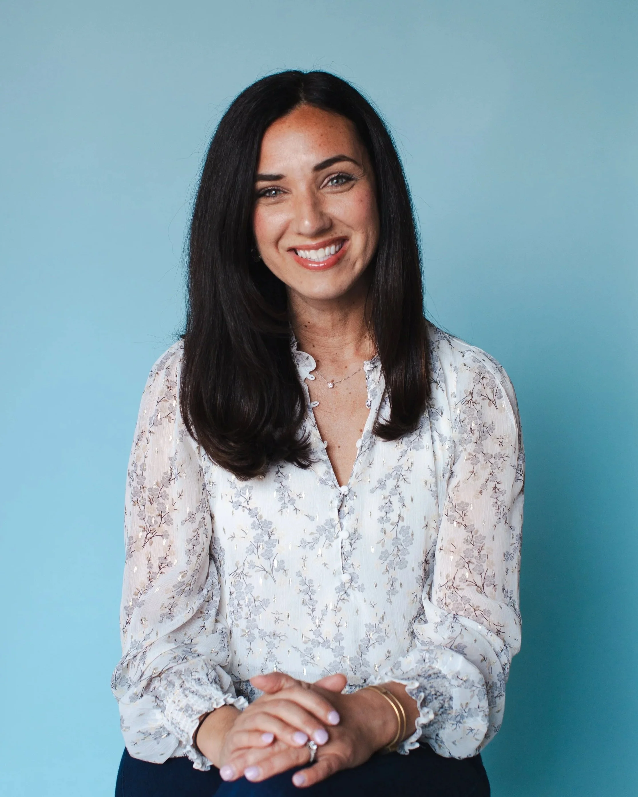 A woman with long dark hair and light skin, smiling, wearing a floral blouse, standing against a light blue background.