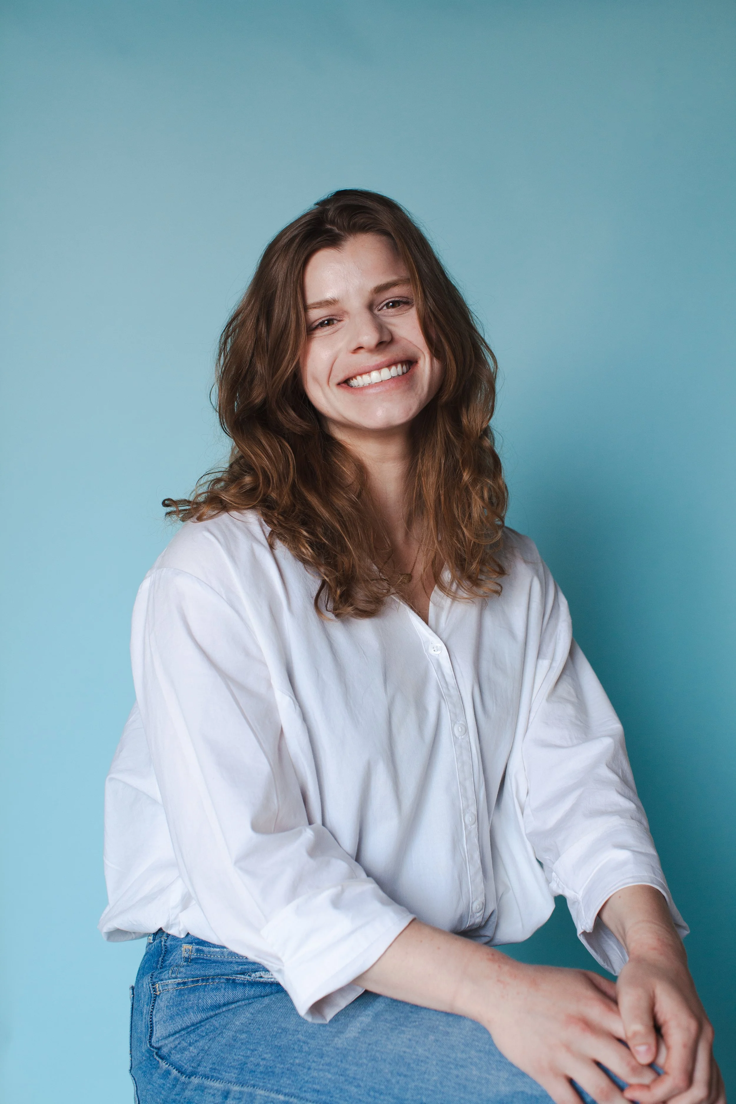 A woman smiling, wearing a white shirt and jeans, sitting against a light blue background.