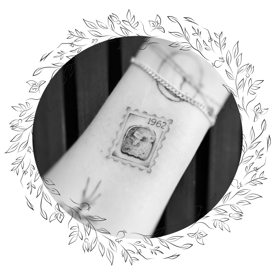 A wrist tattoo of a 1962 postage stamp featuring a profile of a person's head, with a chain bracelet above the tattoo.