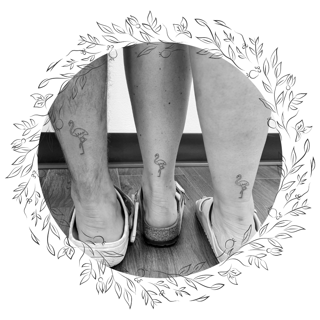 Close-up of a person's lower legs with tattoos of flamingos, wearing sneakers on a wooden floor with a decorative dark overlay.