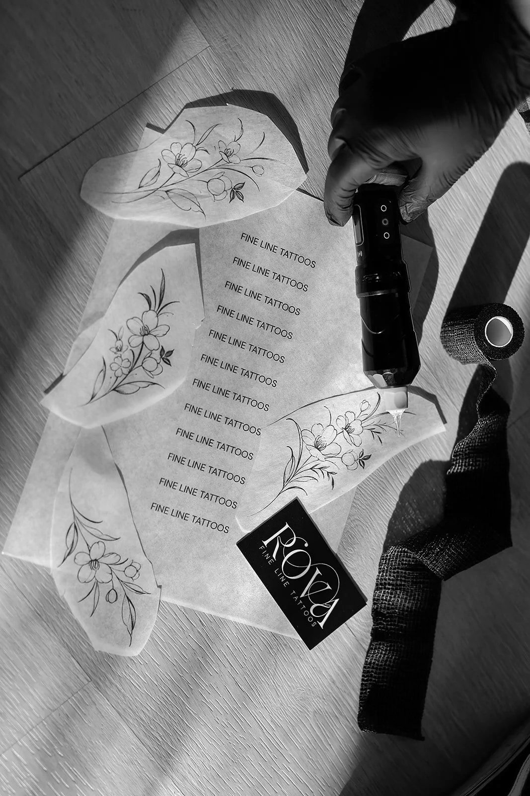 A person wearing black gloves is using a tattoo machine on a design paper with floral tattoo sketches and the words "FINE LINE TATTOOS" repeated multiple times. A roll of black tattoo ink wrap and a business card for "RO FINE LINE TATTOOS" are also visible on the table.