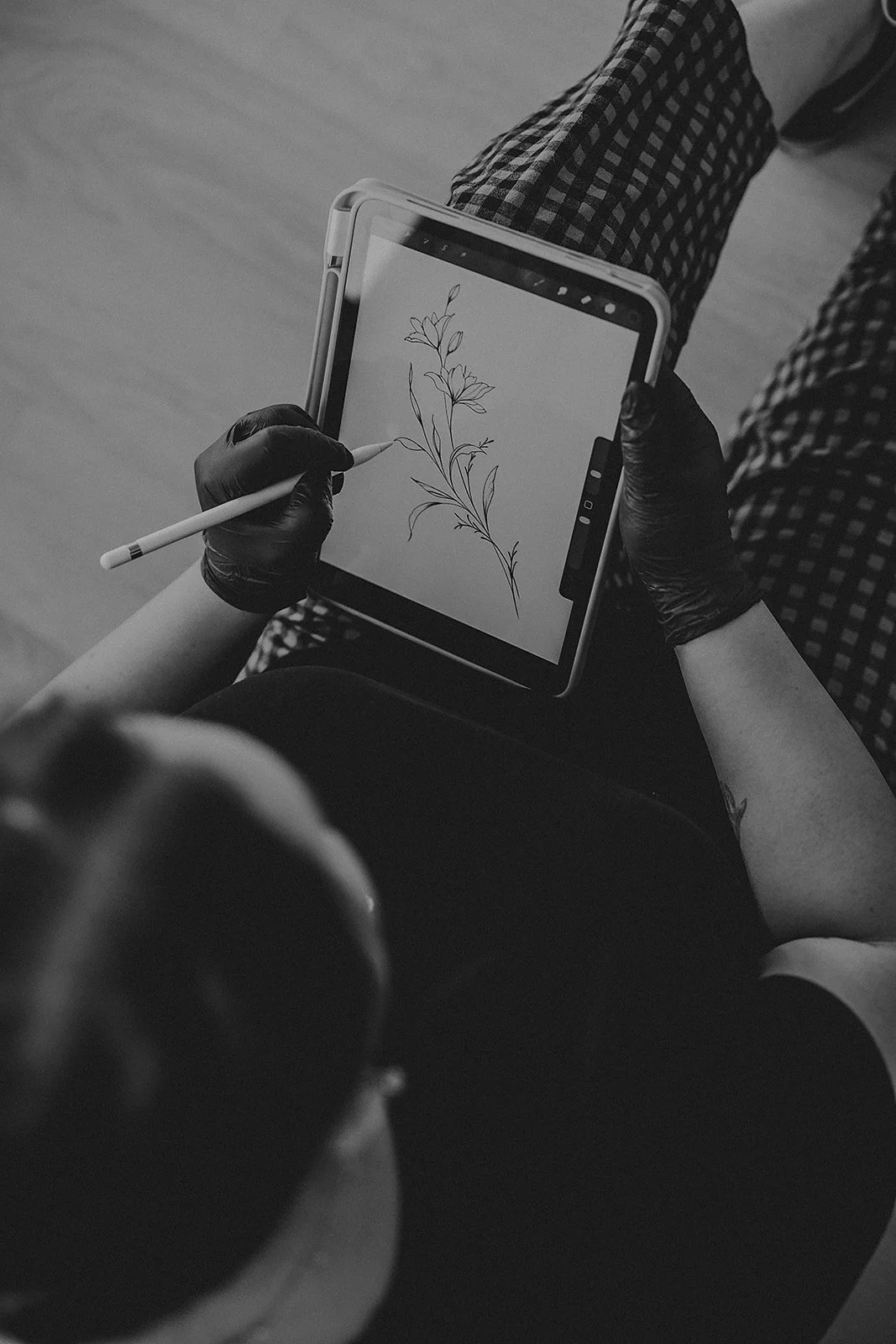 A person with tattoos on their arm, wearing a black glove, is drawing a leafy plant on a tablet screen using a stylus. The person is sitting, wearing checkered pants.