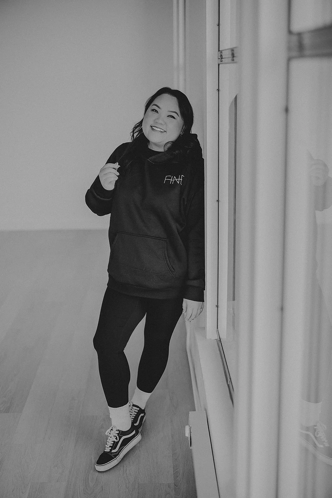 A woman with dark hair smiling and standing by a wall, wearing a hoodie, leggings, sneakers, and socks in black and white.