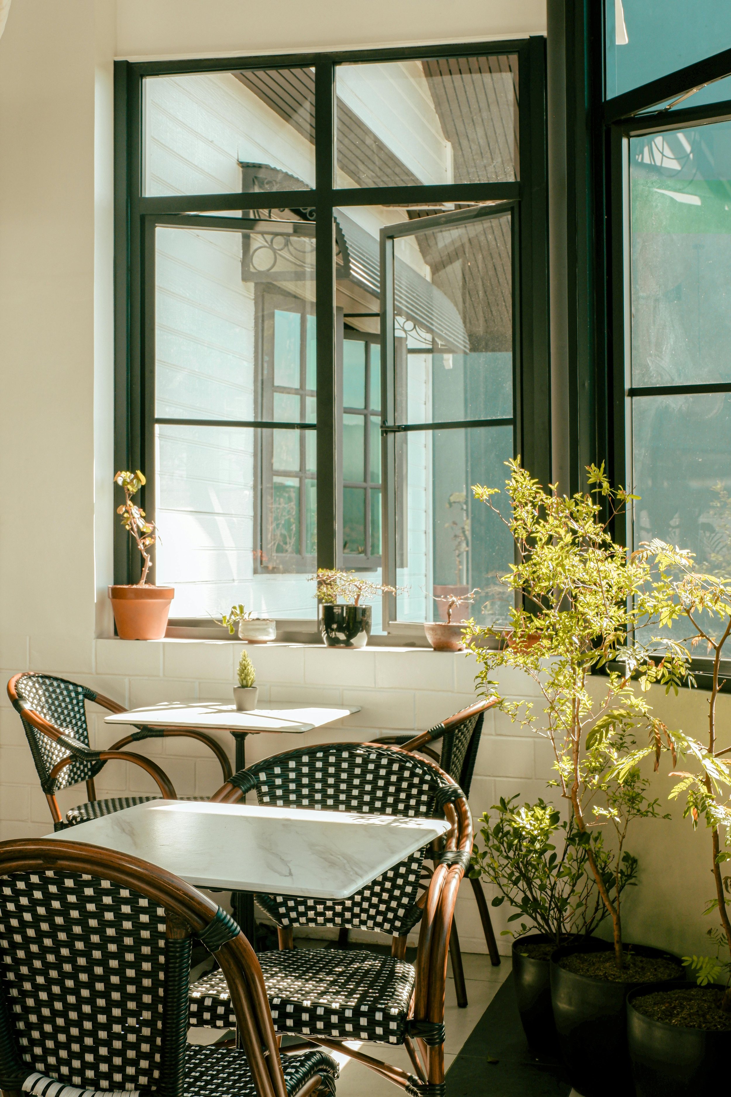 Sunlit cozy indoor space with black and white checkered chairs, white marble tables, and potted plants near open window.