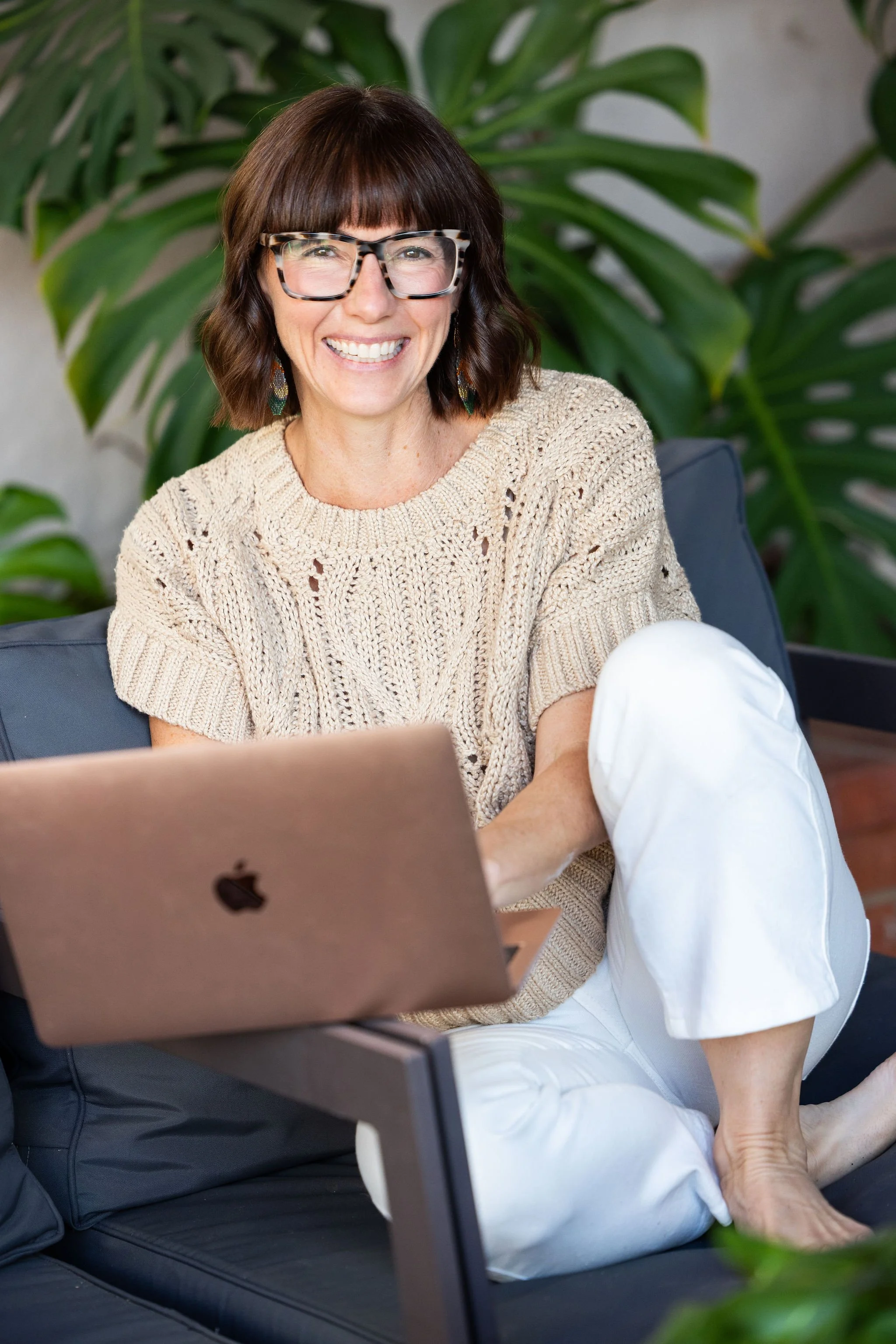 A woman with brown hair, glasses, and earrings sits on a black outdoor sofa, smiling at the camera, with a laptop on her lap and a large green plant behind her.