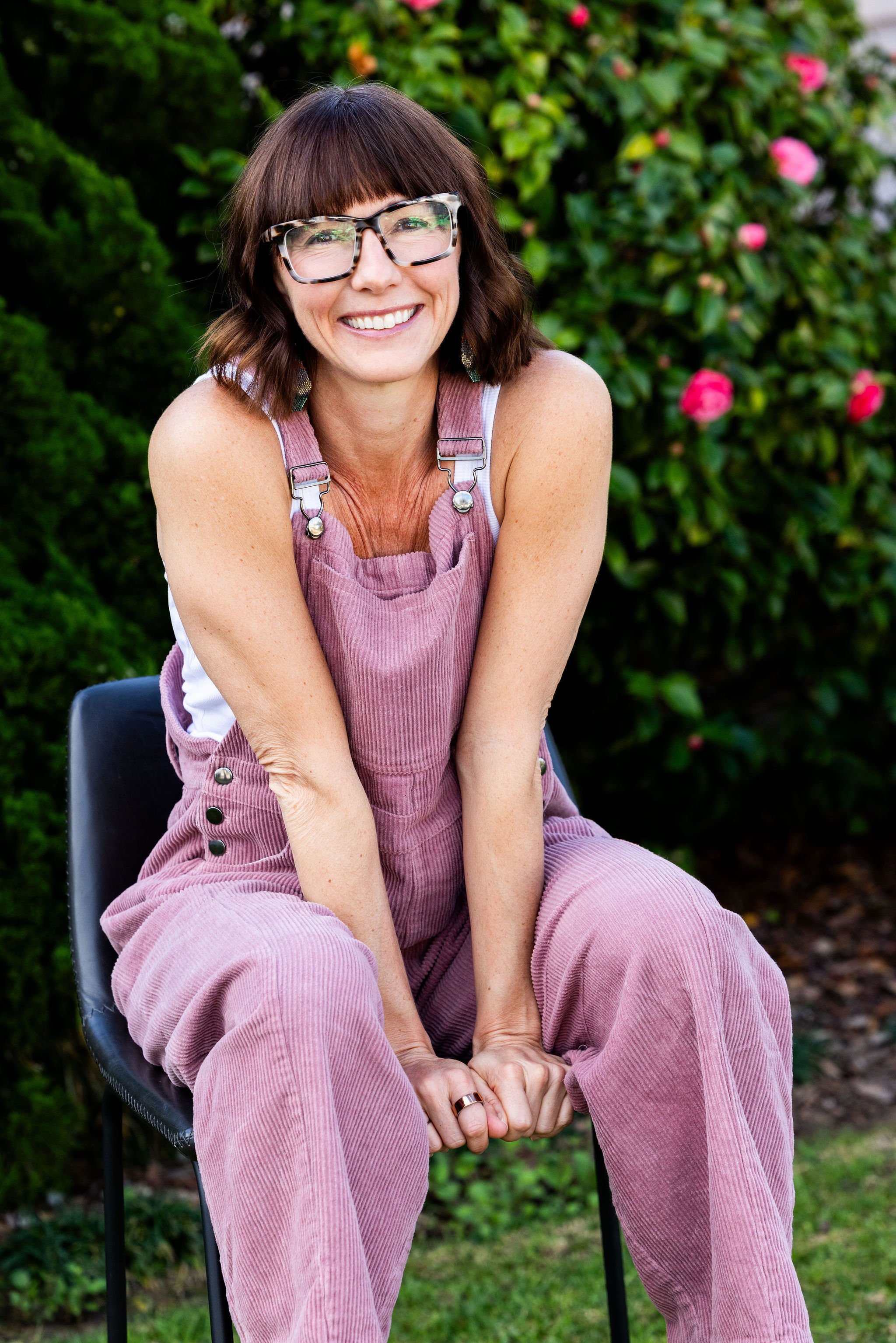 Woman smiling outdoors, wearing glasses and mauve overalls, sitting on a black chair with a green bush and pink flowers in the background.