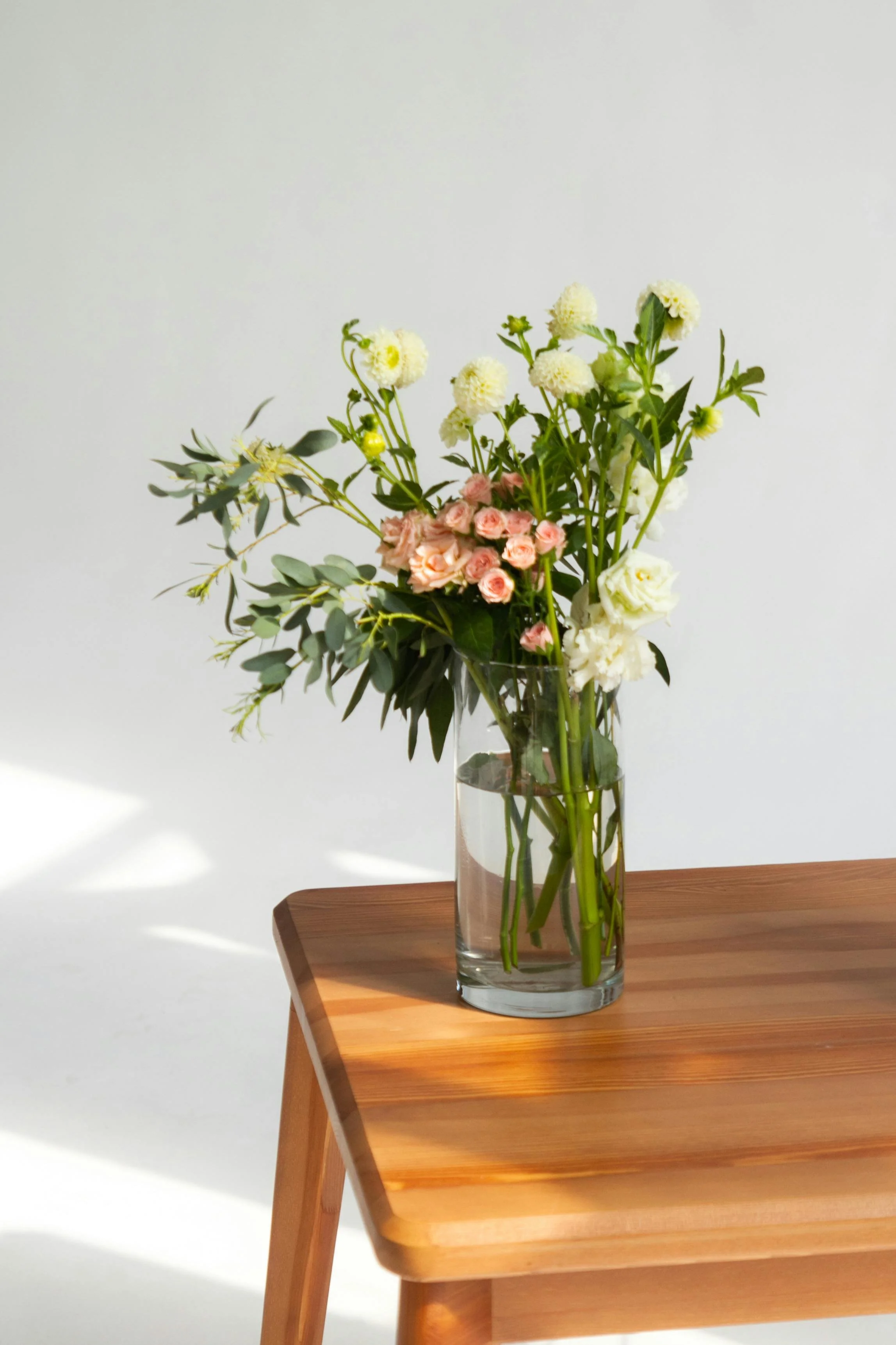 A clear glass vase filled with water holding a bouquet of mixed flowers, including pink roses, white roses, and white pom-pom flowers, placed on a wooden table against a plain white background.