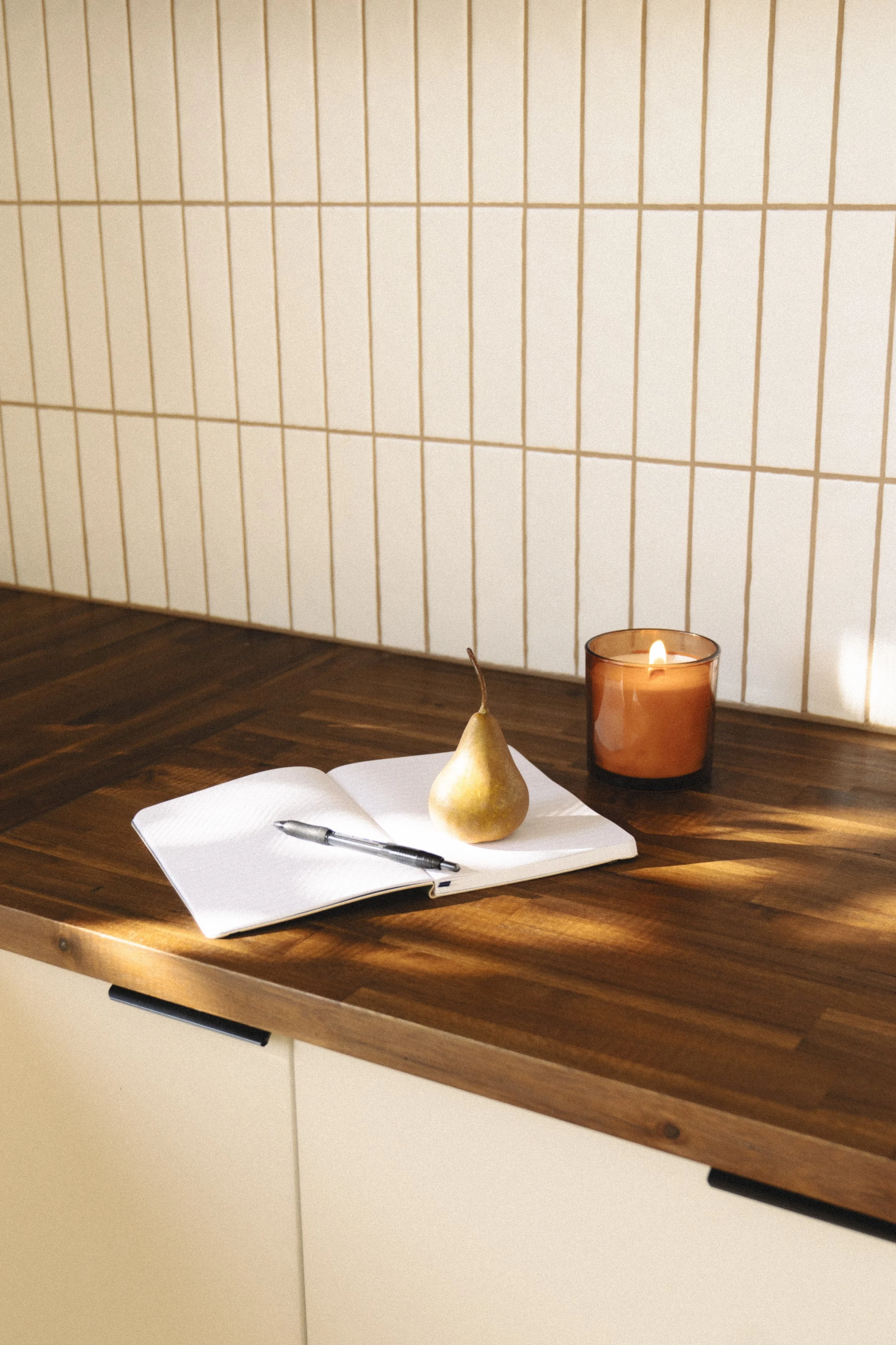 A wooden kitchen countertop with a notebook, a silver pen, a pear, a lit candle in a brown glass holder, and a tiled beige wall in the background.