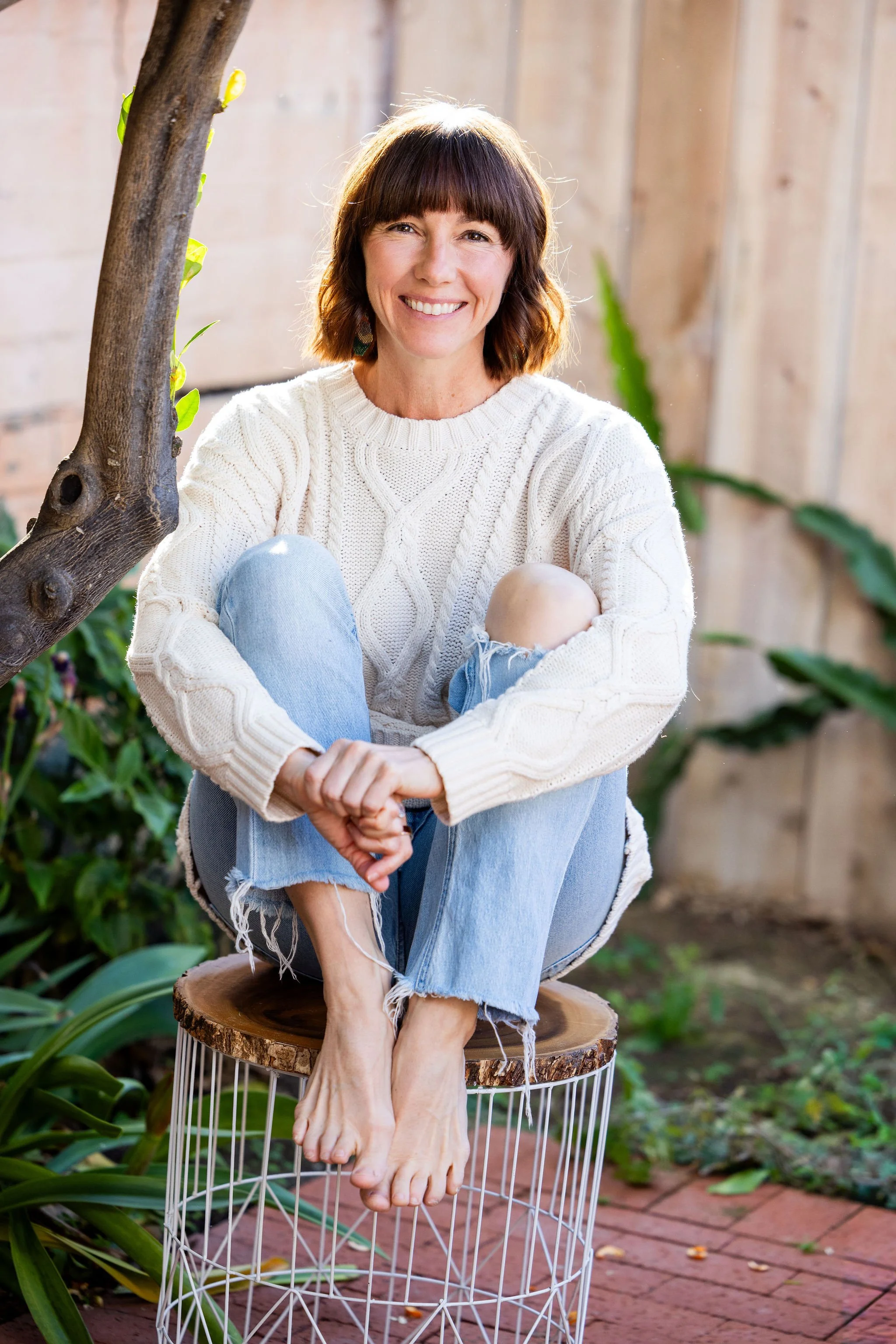 A woman with brown hair and bangs sitting outdoors on a wooden stool without shoes, wearing a cream sweater and ripped jeans, smiling at the camera in a garden setting with brick and wooden fencing.