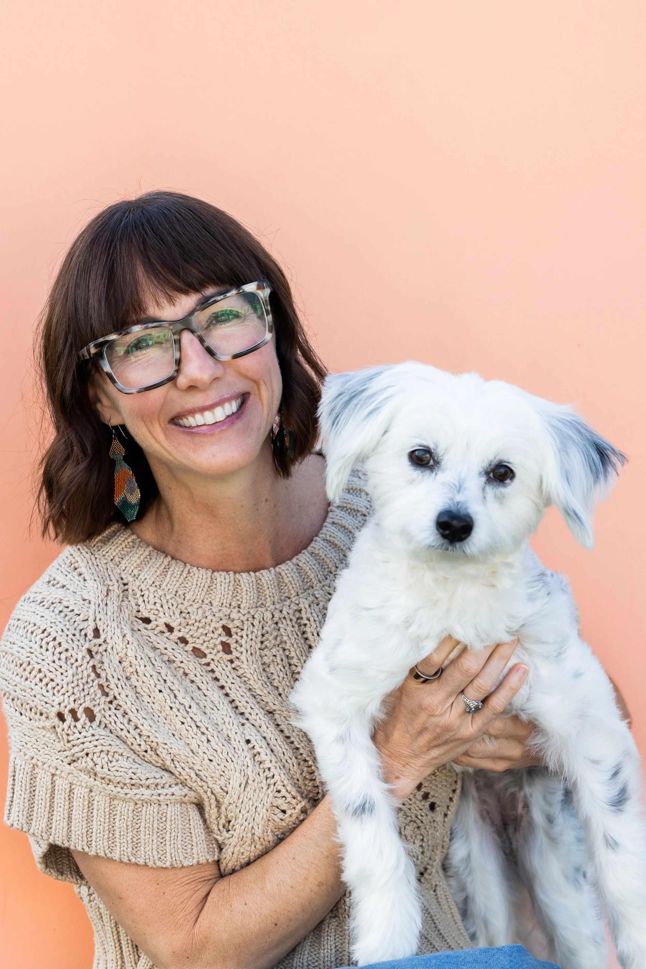 A woman with shoulder-length dark hair, glasses, and earrings, smiling while holding a small white dog with black spots against a peach-colored background.
