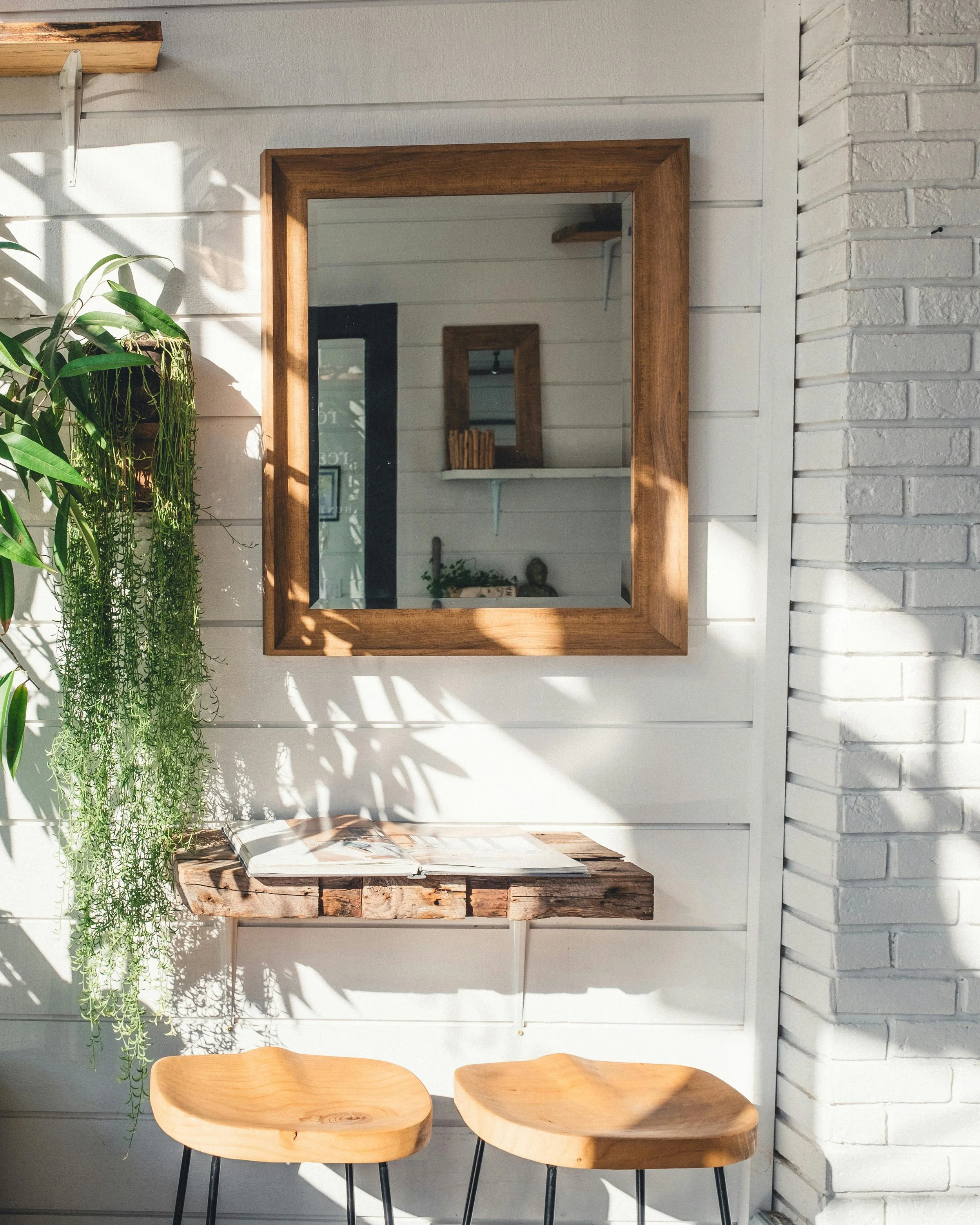 Interior of a cozy, modern space with a white wood-paneled wall, a large wooden framed mirror, a rustic wooden floating shelf with an open magazine, two light wood chairs with black metal legs, a hanging green plant, artificial lighting, and a white brick wall.