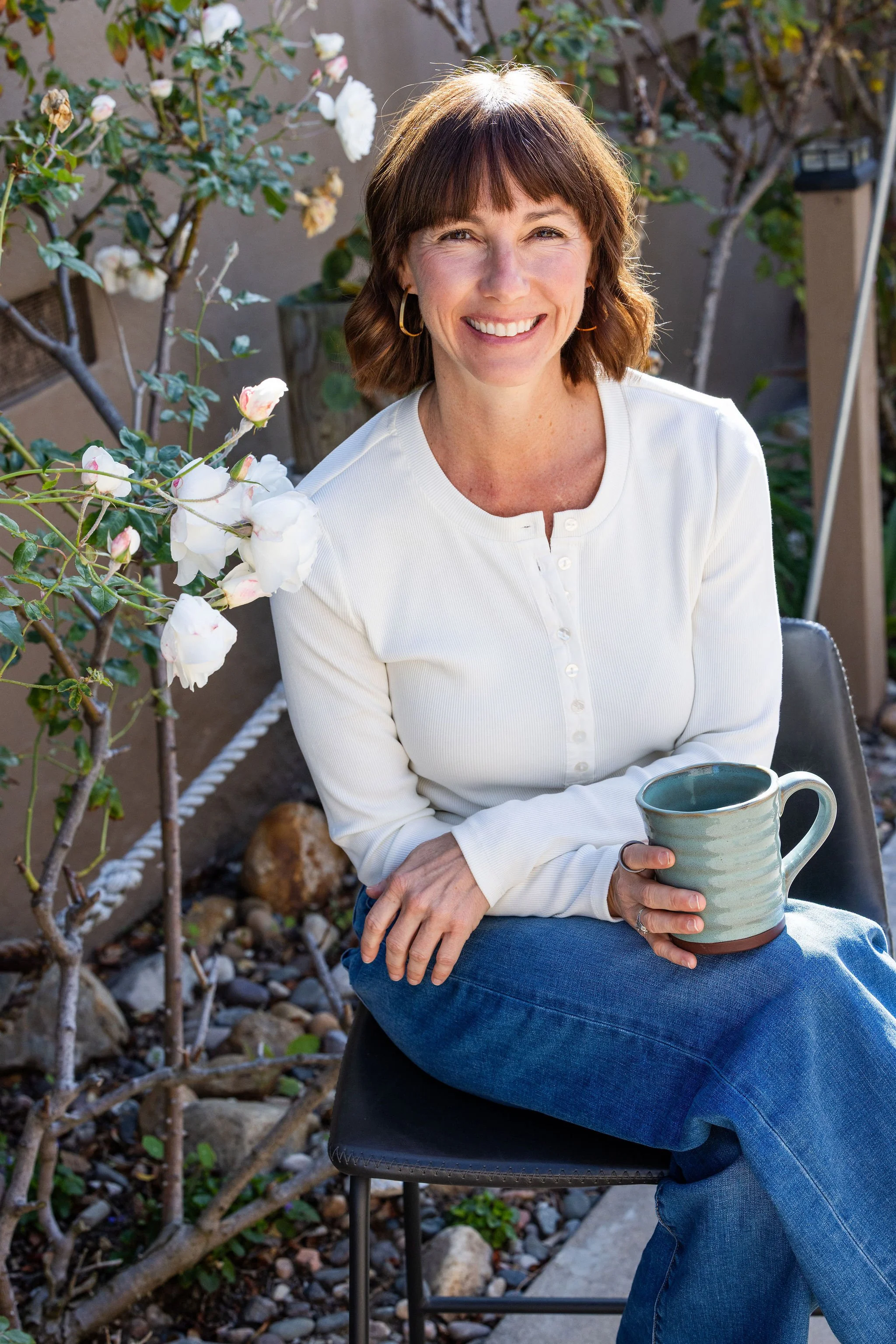 A woman with shoulder-length brown hair and bangs, wearing a white long-sleeve top and blue jeans, sitting on a black chair outdoors next to a flowering plant with white flowers. She is smiling and holding a ceramic mug.
