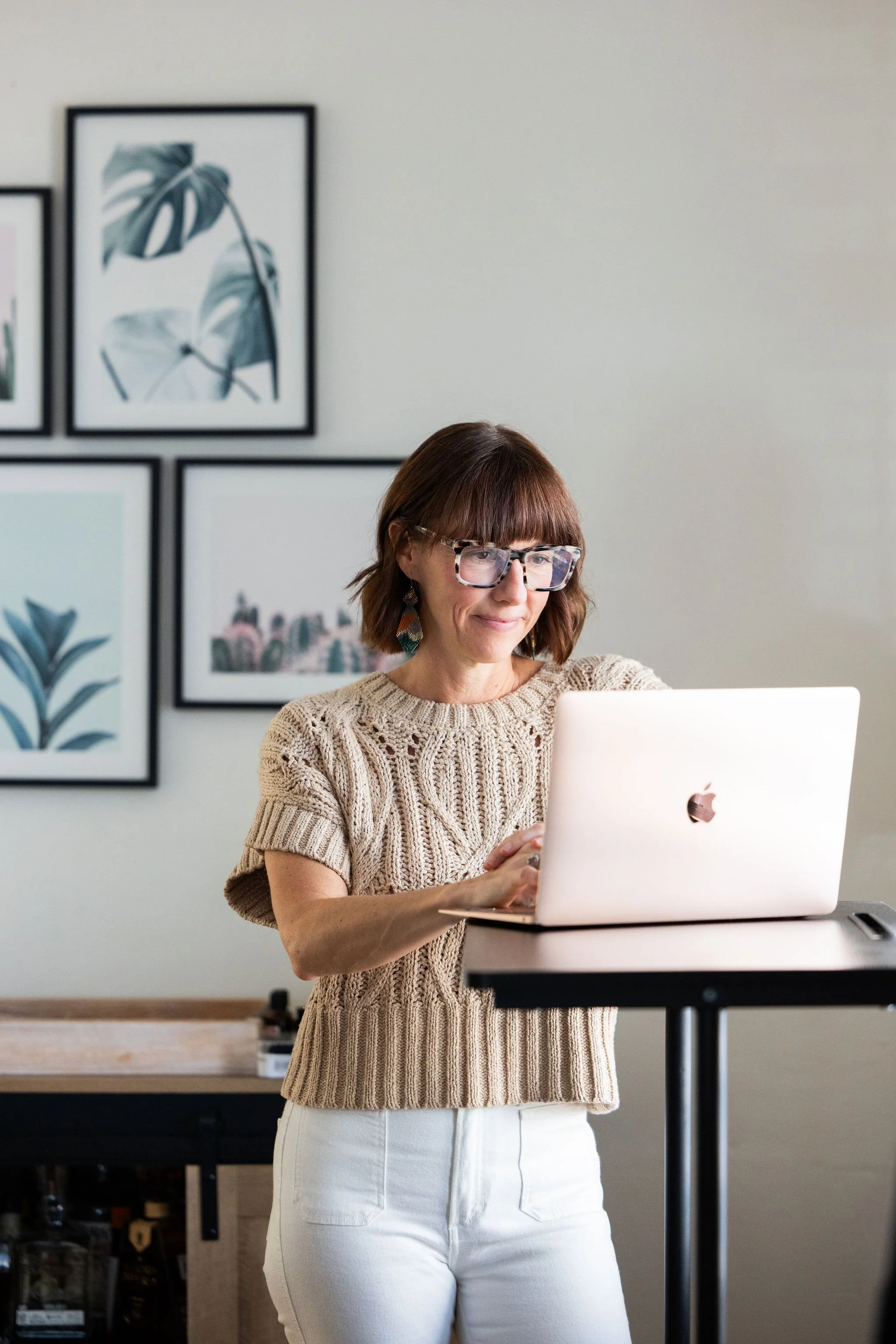 A woman with short brown hair, glasses, and earrings standing at a high table using a white Apple MacBook, in a room with framed botanical artwork on the wall.