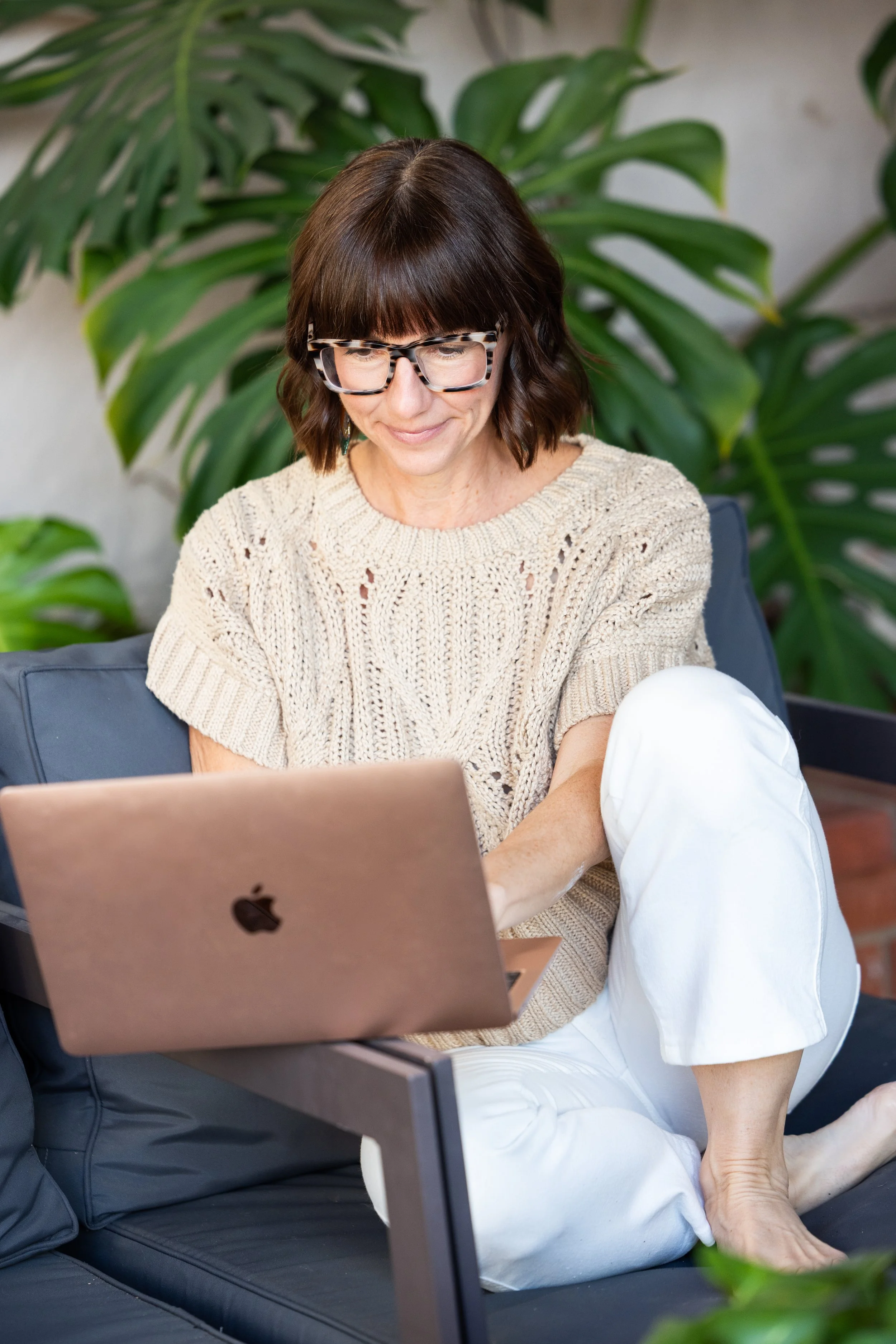 A woman with short brown hair, glasses, wearing a beige knitted sweater and white pants, sitting on a black couch outdoors with large green plants in the background, working on a MacBook.