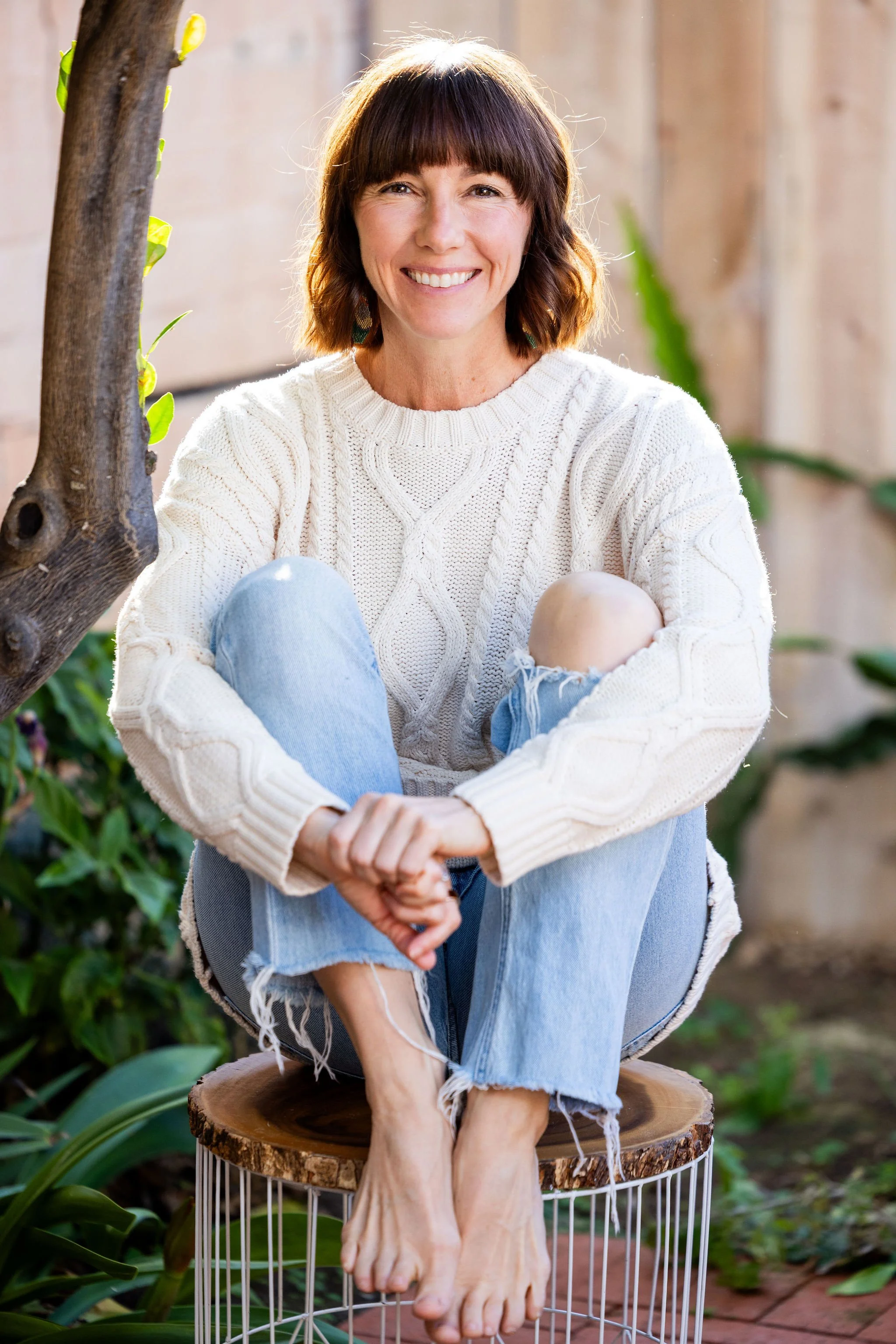 A woman with short brown hair and a white sweater, sitting on a wooden stool outdoors, smiling with her legs crossed and barefoot, surrounded by greenery.
