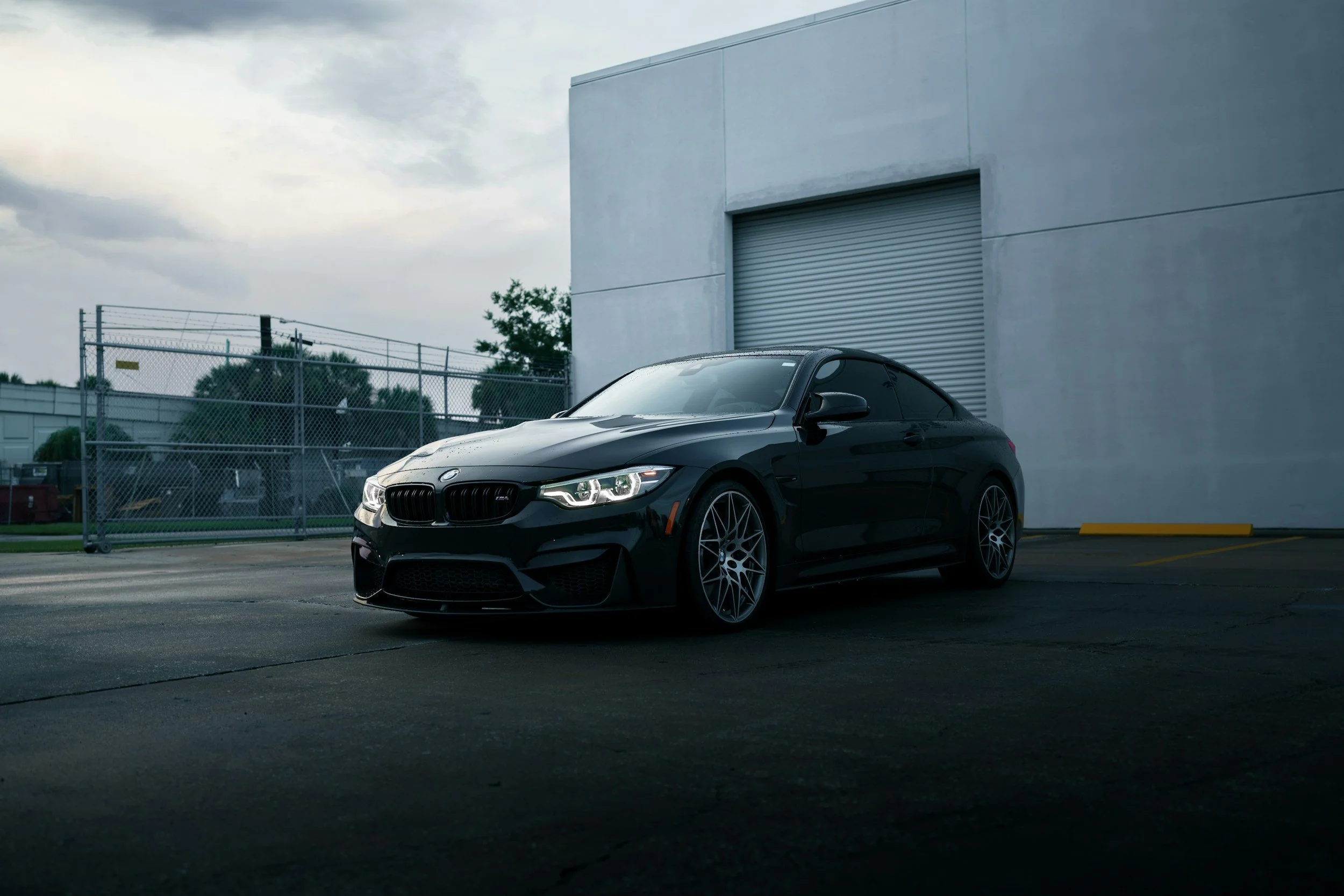 A black BMW coupe parked on a paved lot near a large white building with a rolling shutter door.