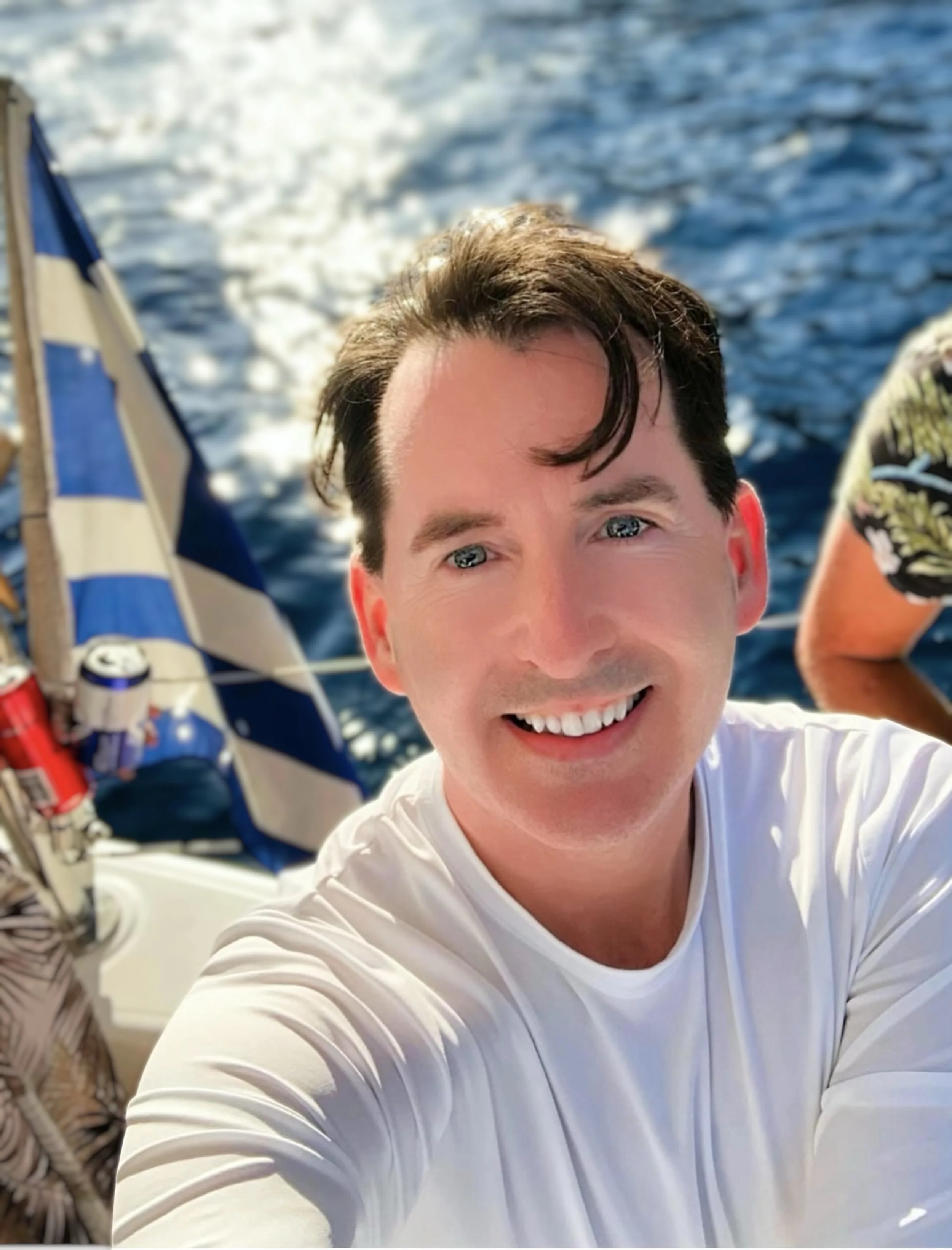 A non-binary person smiling and taking a selfie on a catamaran in Santorini with a view of the sea in the background. There is a blue and white striped Greek flag, and the glare of the sun on the sea visible from the boat.