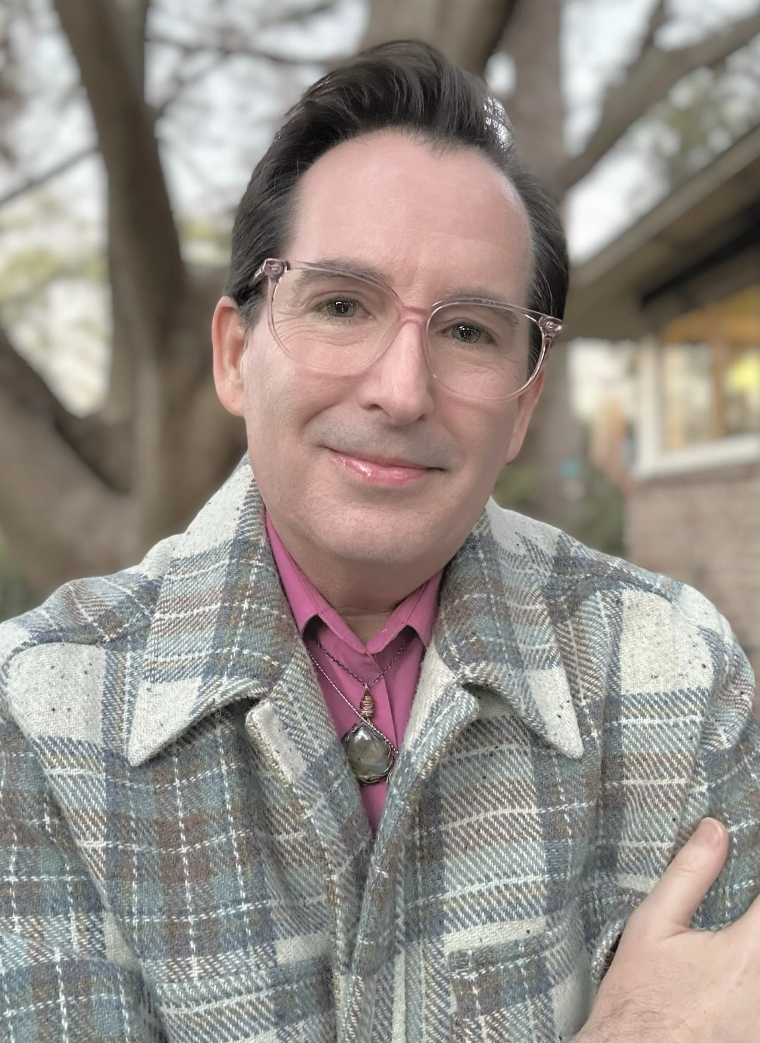 A non-binary person with glasses, dark hair, and a smile, wearing a patterned jacket over a pink shirt, standing outdoors with a tree and house in the background.