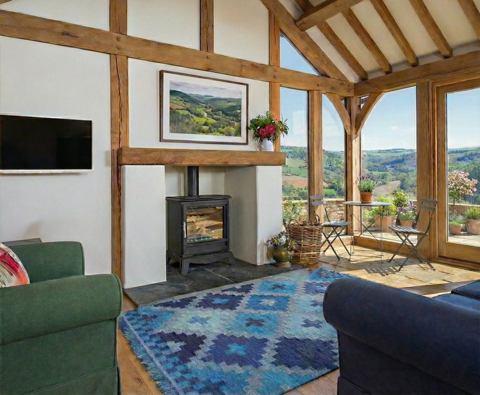 Living room with green and black sofas, a colorful blue patterned rug, a wood stove, and large windows overlooking a scenic countryside landscape with rolling hills and potted plants outside.