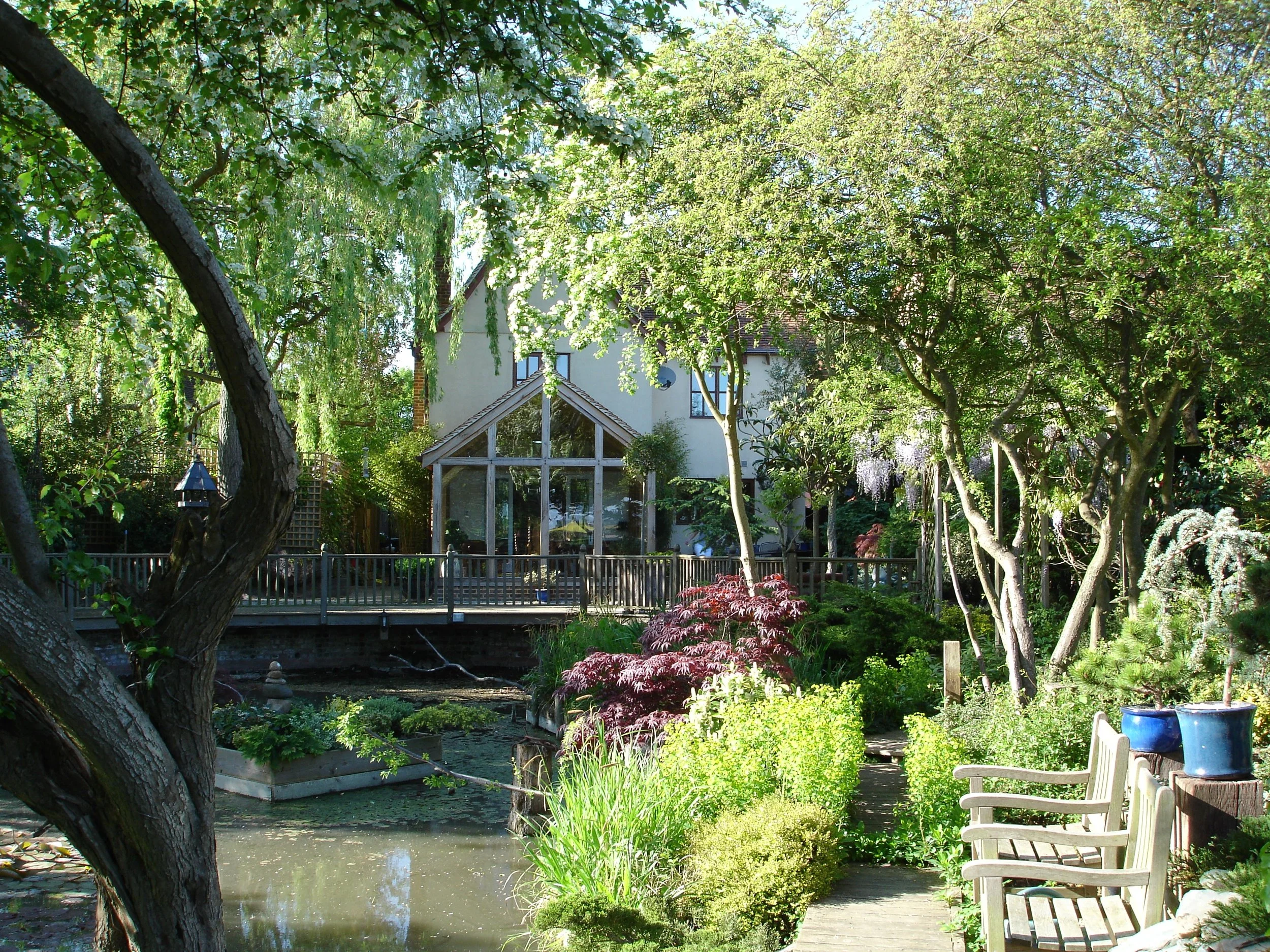 A lush garden with a pond, trees, bushes, and a wooden pathway. A house with a glass extension is visible in the background.