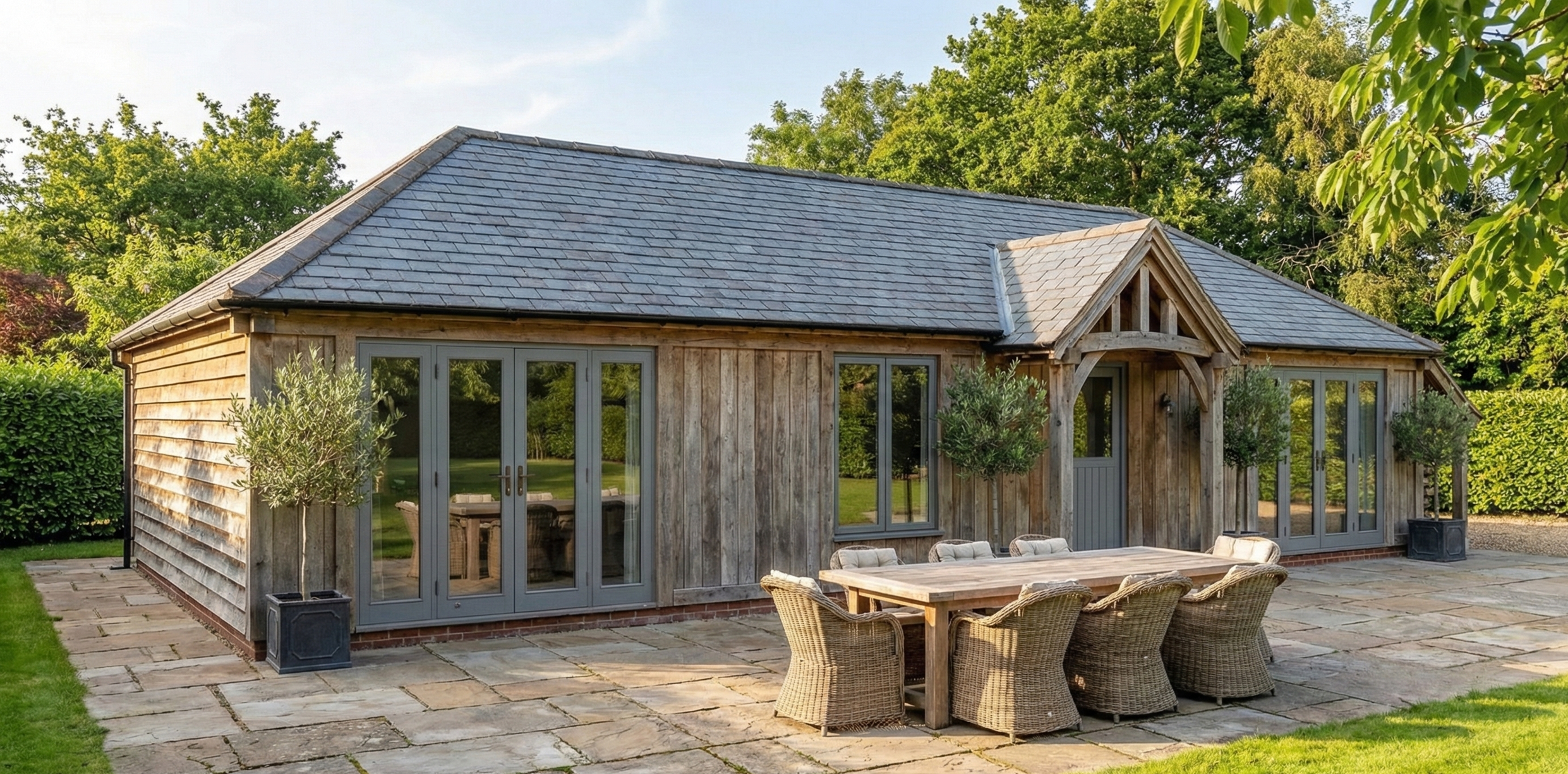 A rustic wooden house with large glass doors and windows, surrounded by green trees and bushes, with a stone patio and outdoor dining area with a wooden table and wicker chairs.