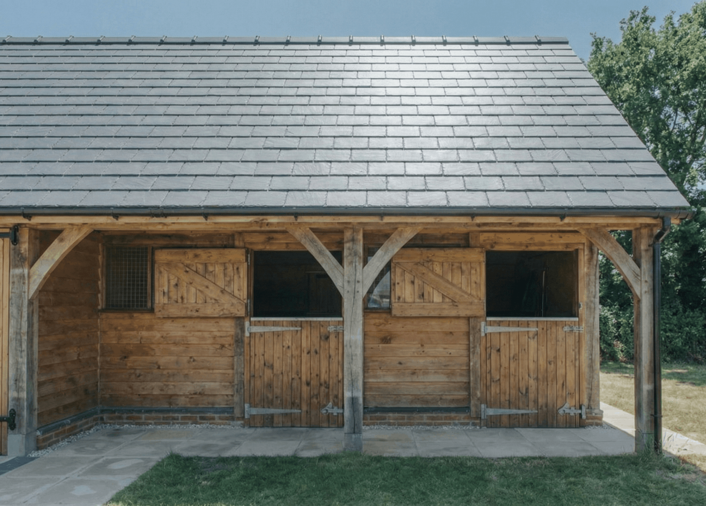 Wooden horse stable with double doors, small barred window, and a gray shingled roof, surrounded by grass and trees.