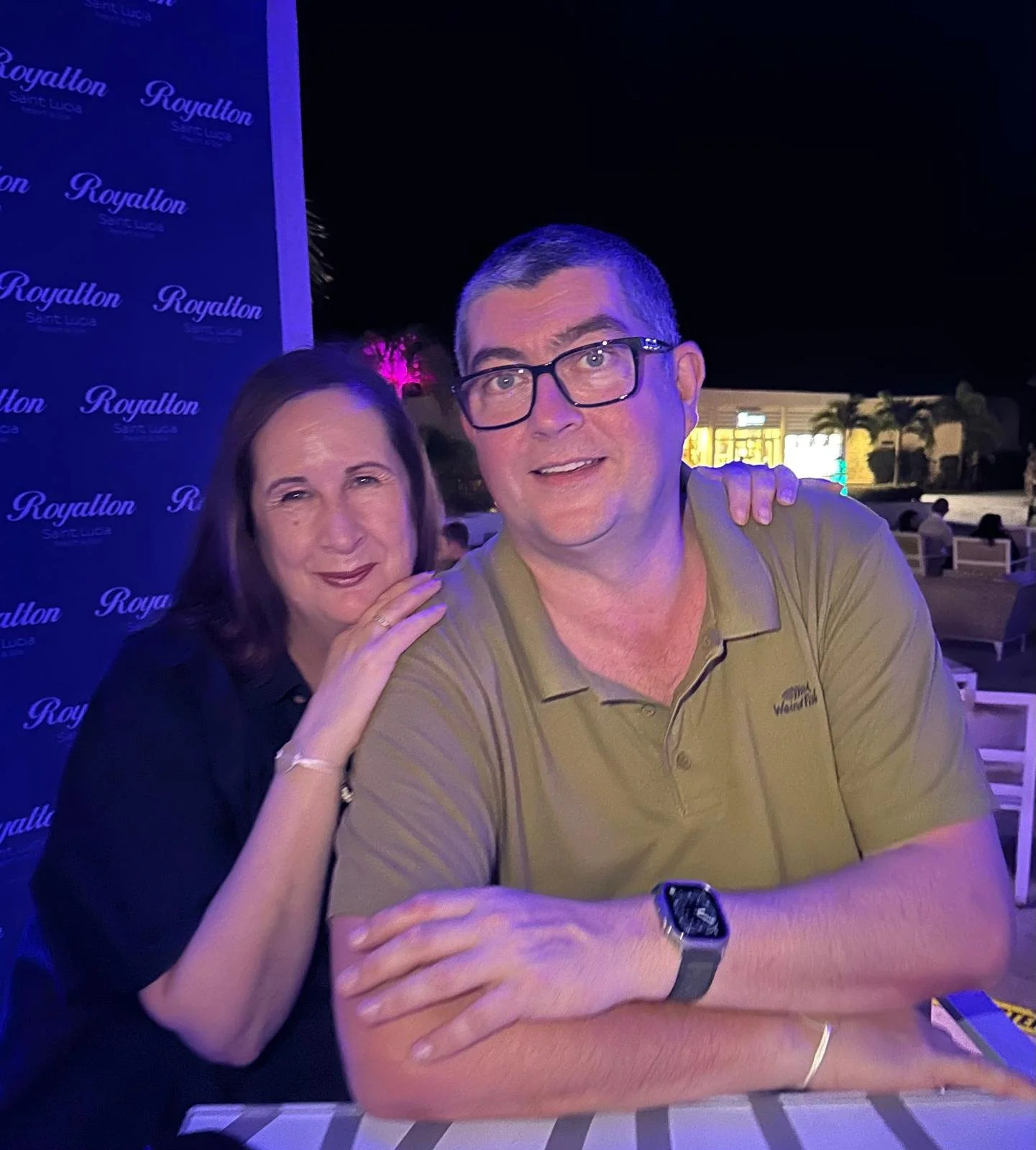 A woman and a man posing together at an outdoor venue at night. The woman has brown hair, wearing a black top, and resting her hand on the man's shoulder. The man has short gray hair, glasses, and is wearing a tan polo shirt with a watch. There is a blue backdrop with the text "Royallon Saint Lucia" on it, and in the background, there are palm trees and buildings illuminated at night.