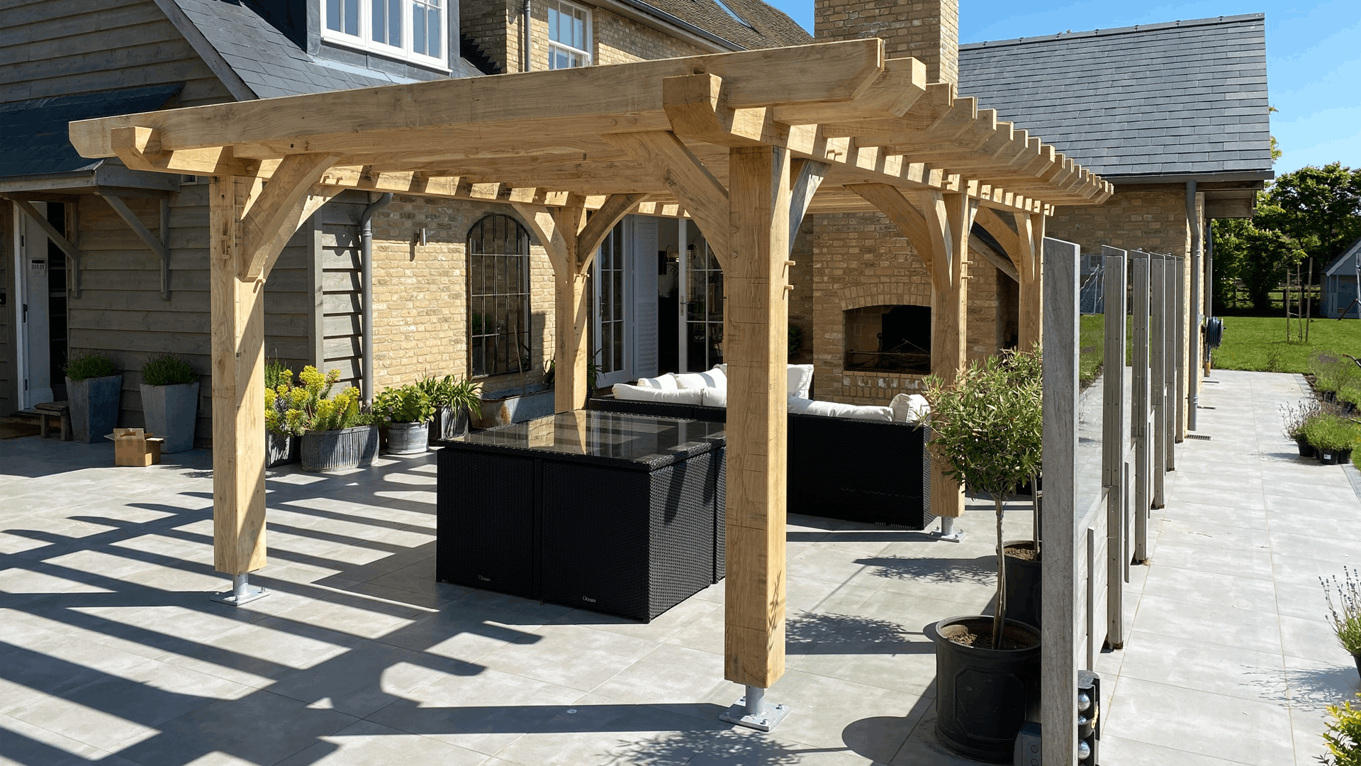 A backyard patio with a wooden pergola, outdoor seating, potted plants, and a brick house with large windows and an outdoor fireplace.