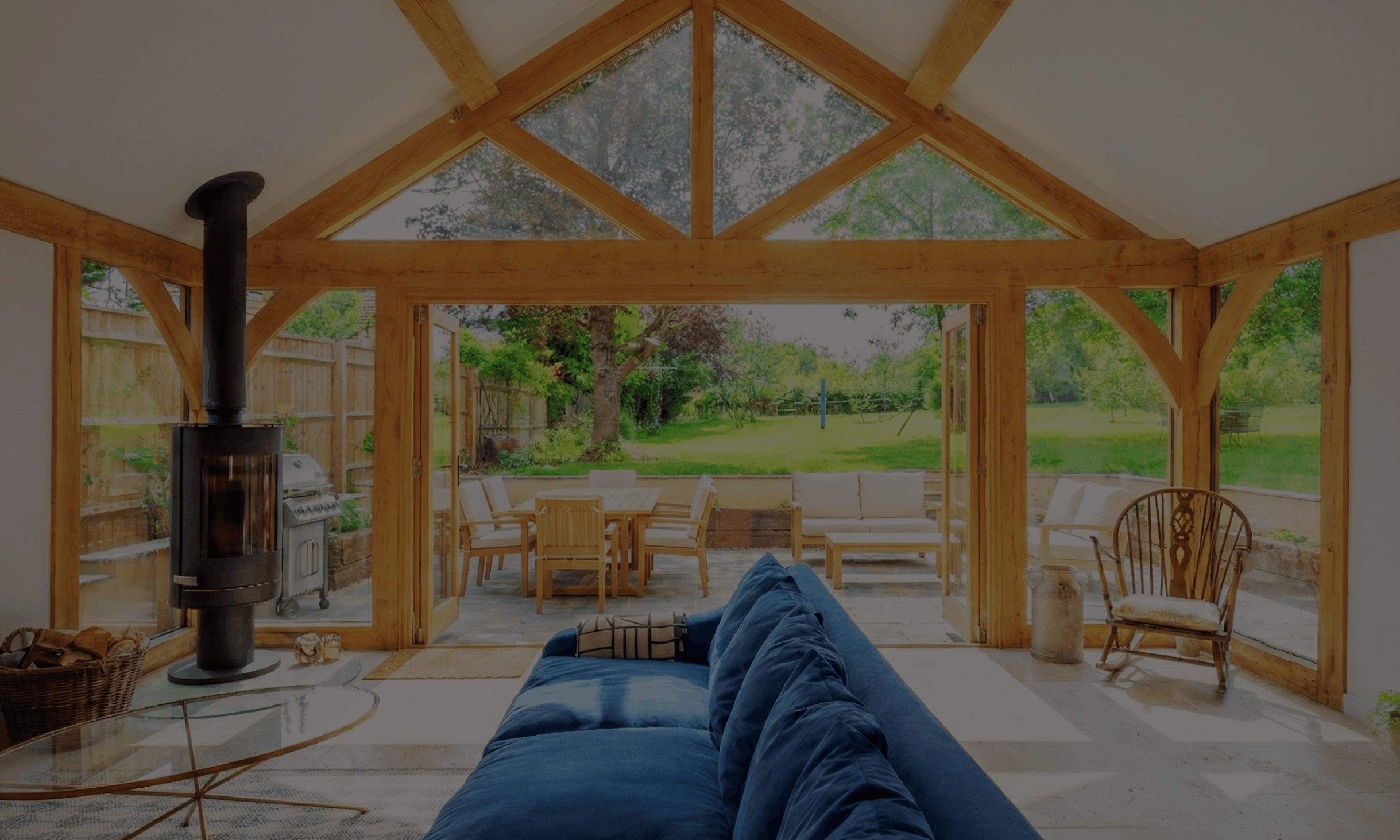 Interior view of a sunroom with wooden beams, a blue sofa, a glass coffee table, and a view of a lush backyard with outdoor furniture and green trees.