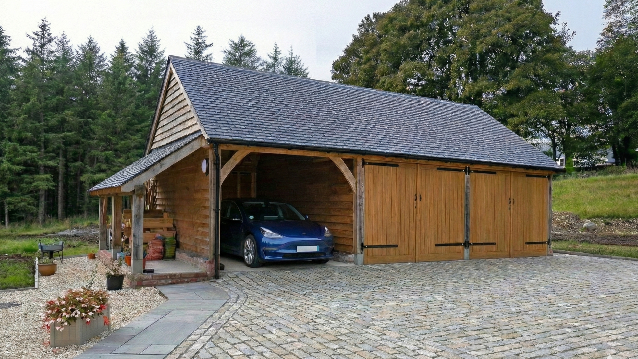 A wooden garage with a pitched roof, partially open, containing a blue electric car parked inside. The garage is surrounded by a paved driveway and landscaped garden with potted plants, trees, and a gravel area.