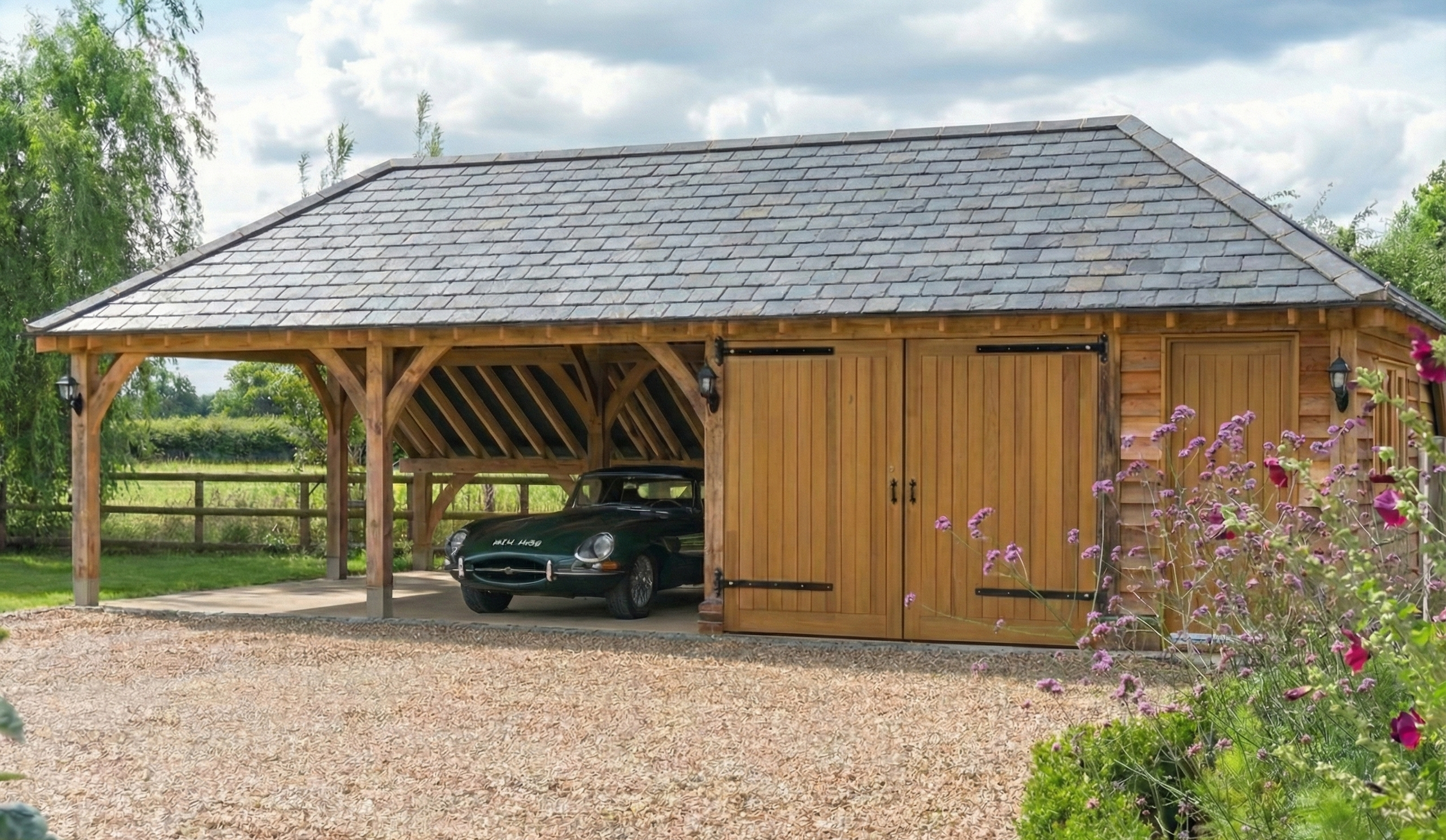 A wooden garage with a car parked inside, surrounded by greenery and pink flowers, under a cloudy sky.